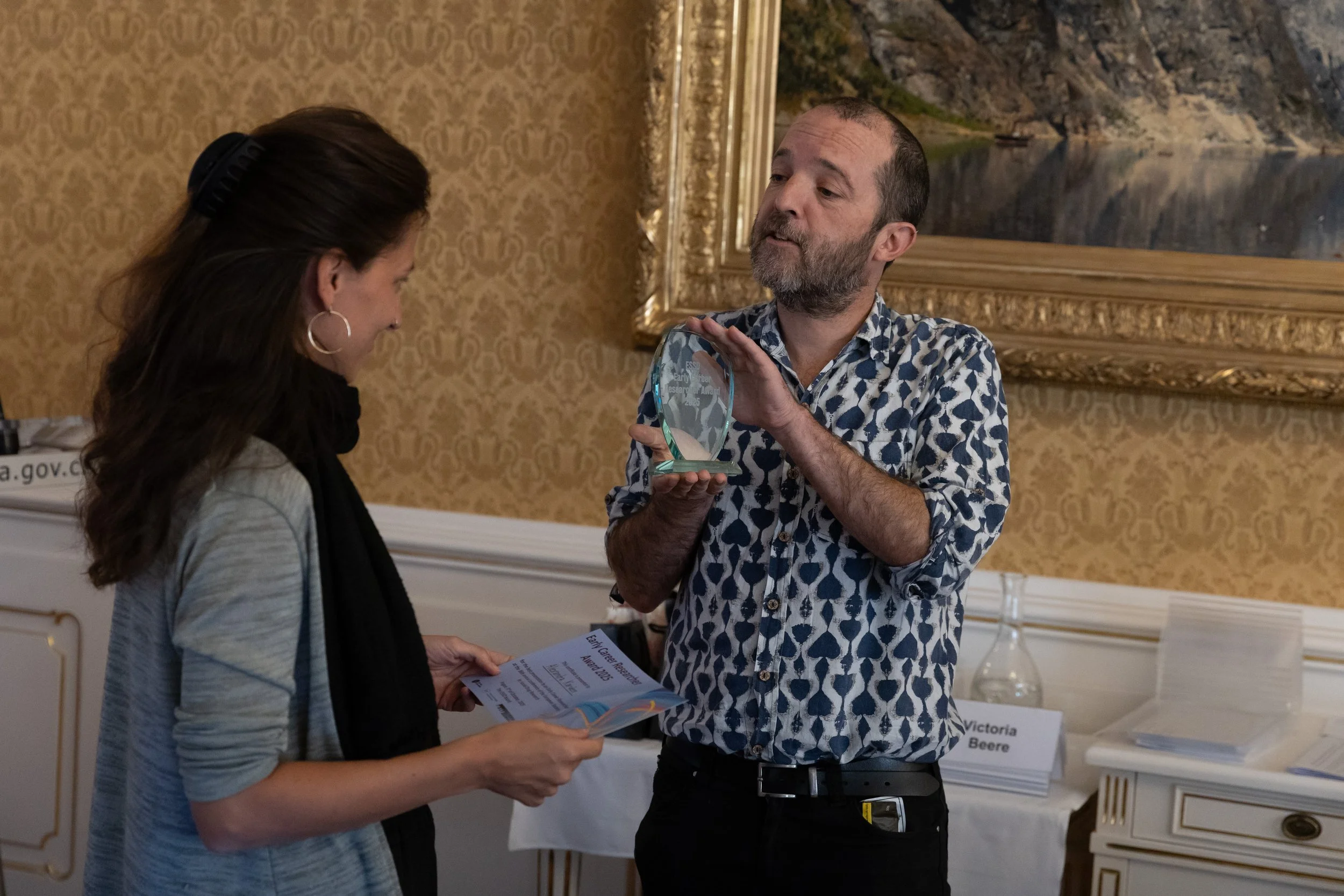 A man with a beard in a patterned shirt is giving an award to a woman with long dark hair, hoop earrings, and a gray sweater with a black scarf. They are indoors with a large framed painting in the background.