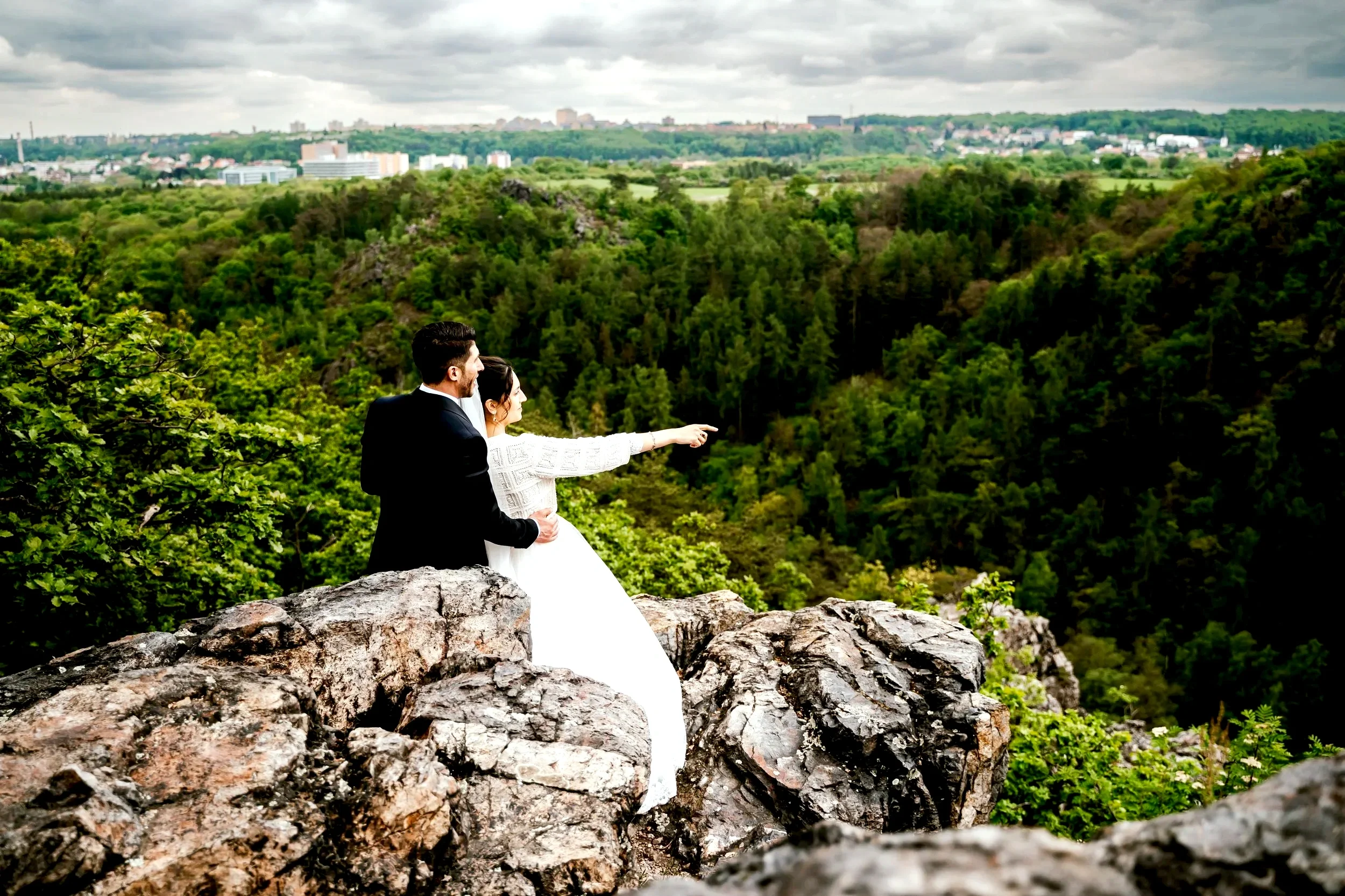 A bride and groom in wedding attire sitting on rocks, overlooking a lush green forest and cityscape in the distance, with cloudy sky overhead.