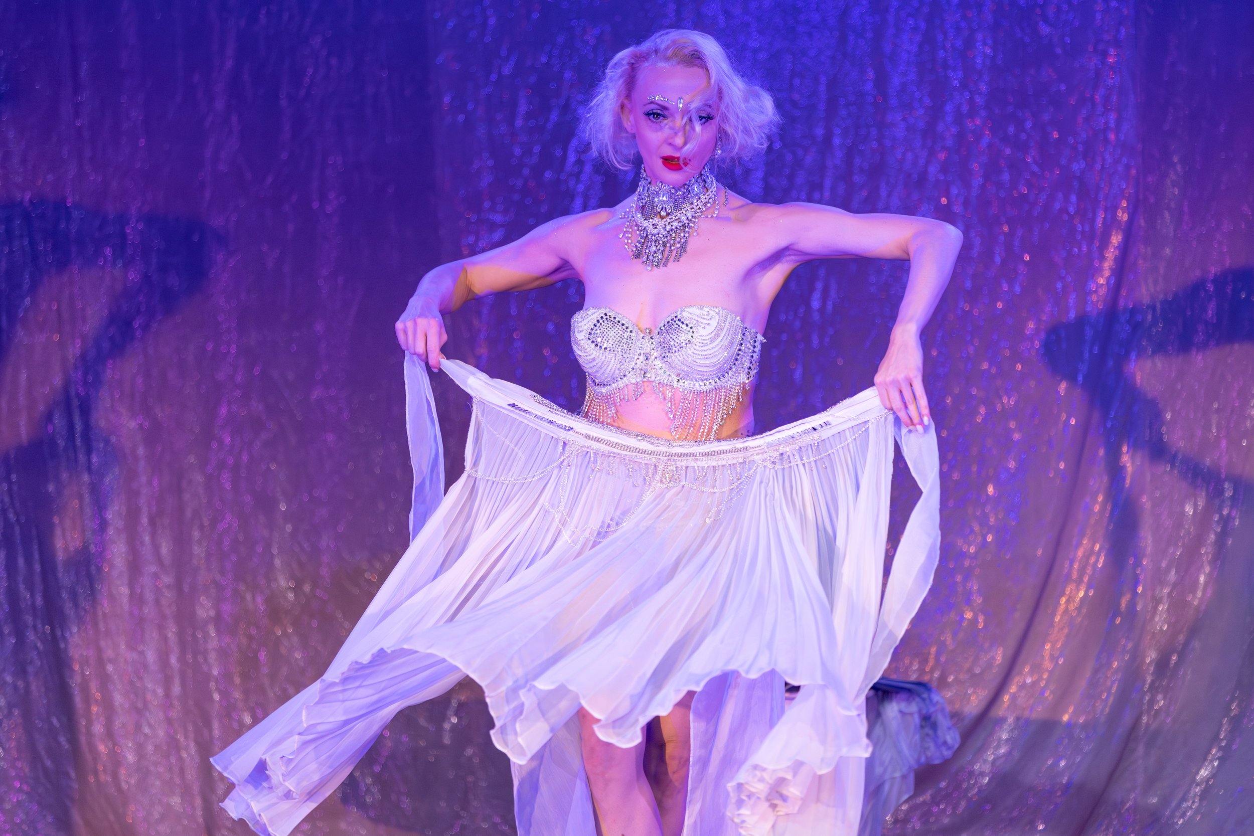 A woman dressed in a shimmering, ornate bra and flowing white skirt, holding the skirt out in a pose, standing in front of a purple curtain backdrop.