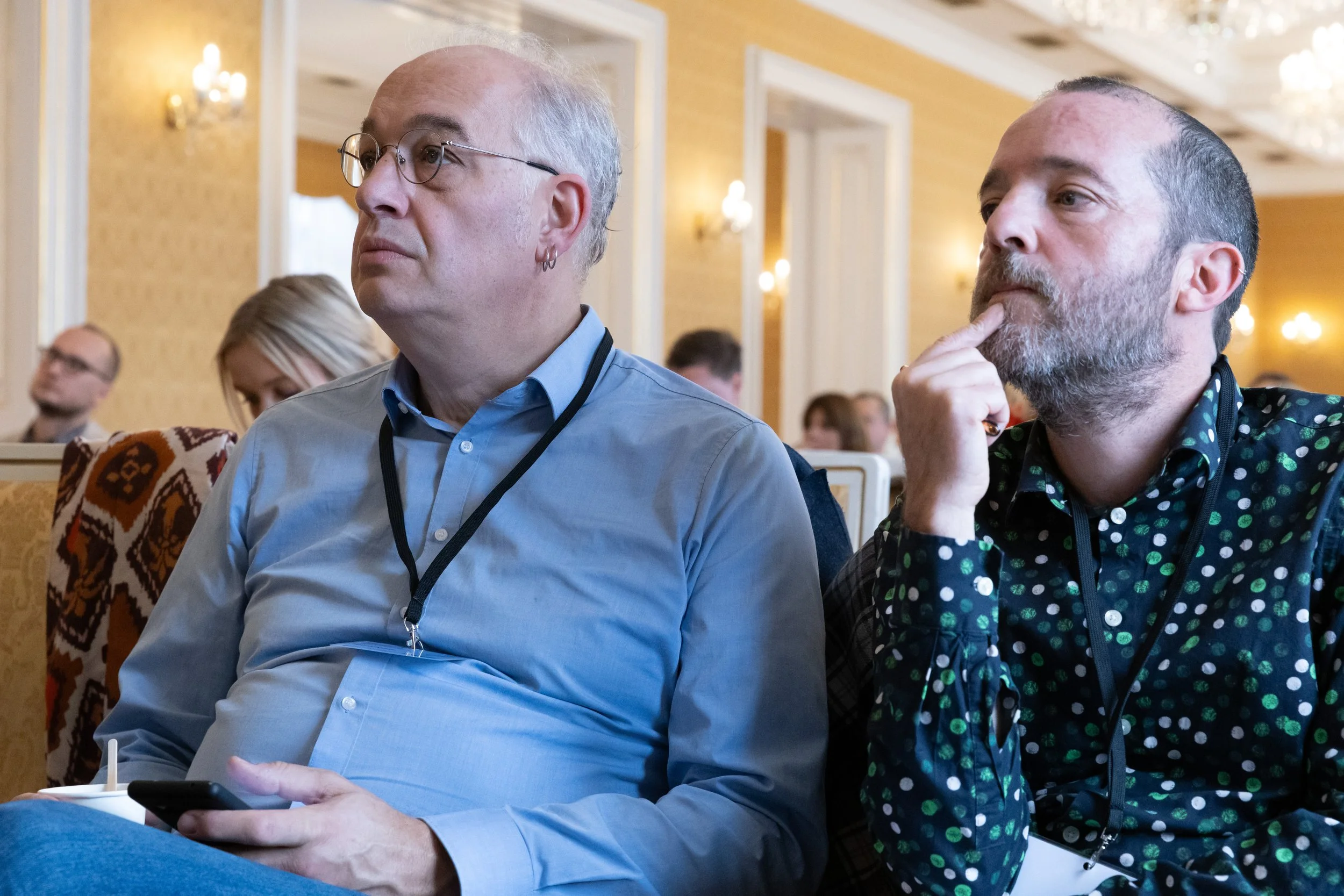 Two men are sitting in an ornate conference room, attentively listening during a presentation or lecture, with other attendees visible in the background.