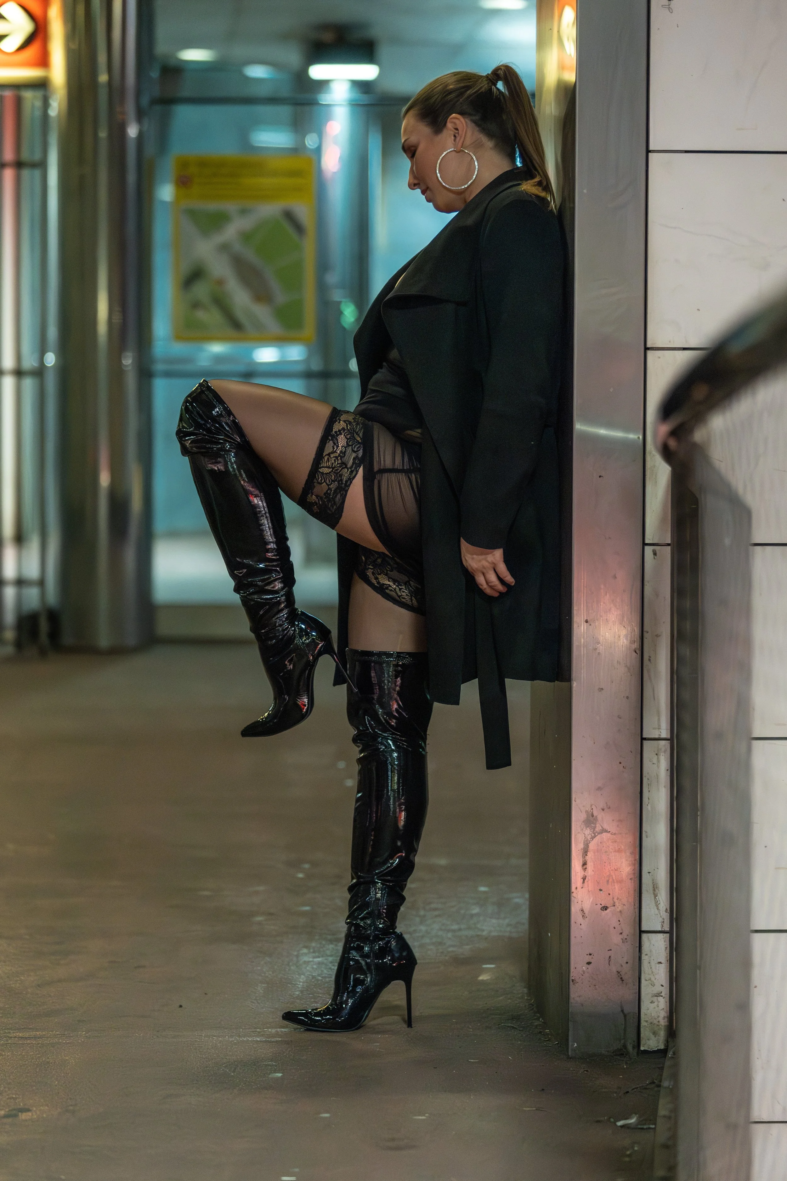 A woman in black clothing and thigh-high shiny black boots stands against a wall, with her left leg raised and foot resting on the wall, in a subway or train station.