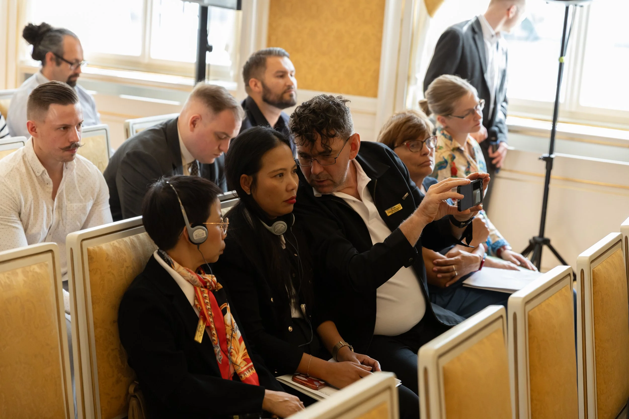 Group of people seated in rows at a conference, some wearing headphones, with a man showing something on his phone to a woman in the front row.