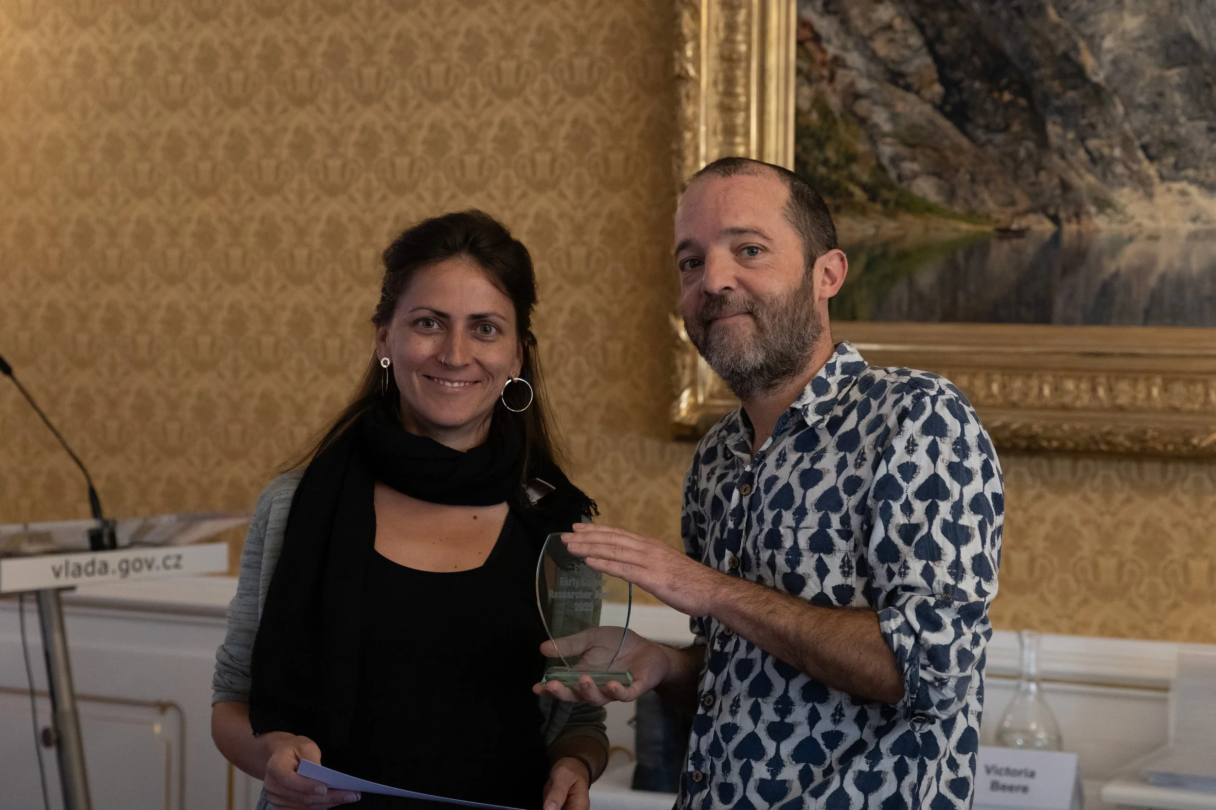 A woman and a man standing indoors, smiling, holding an award or plaque together. The woman has dark hair, is wearing a black top, gray cardigan, and large hoop earrings. The man has short hair, a beard, and is wearing a patterned shirt. There is a l