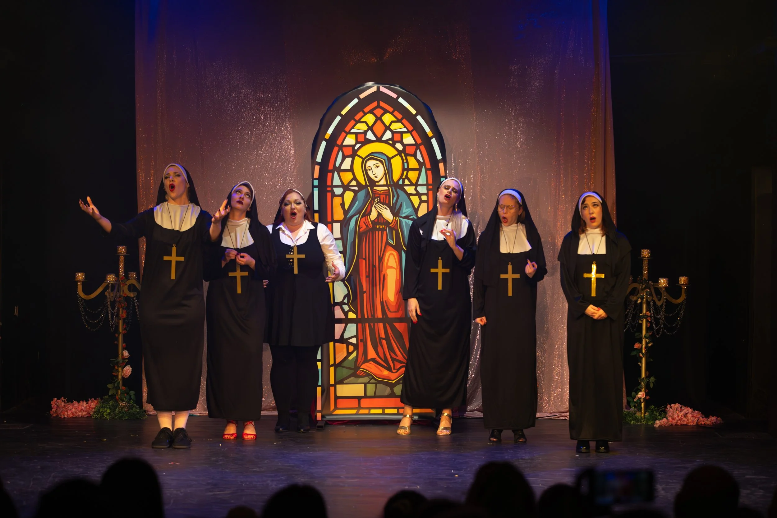 Six women dressed as nuns singing on stage with a stained glass window background