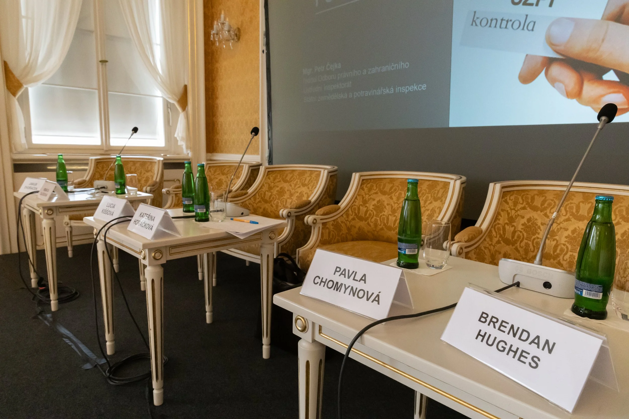 Conference room setup with ornate chairs behind tables, each with a nameplate, green glass bottles of water, glasses, and microphones. Large screen at the front shows a presentation slide.