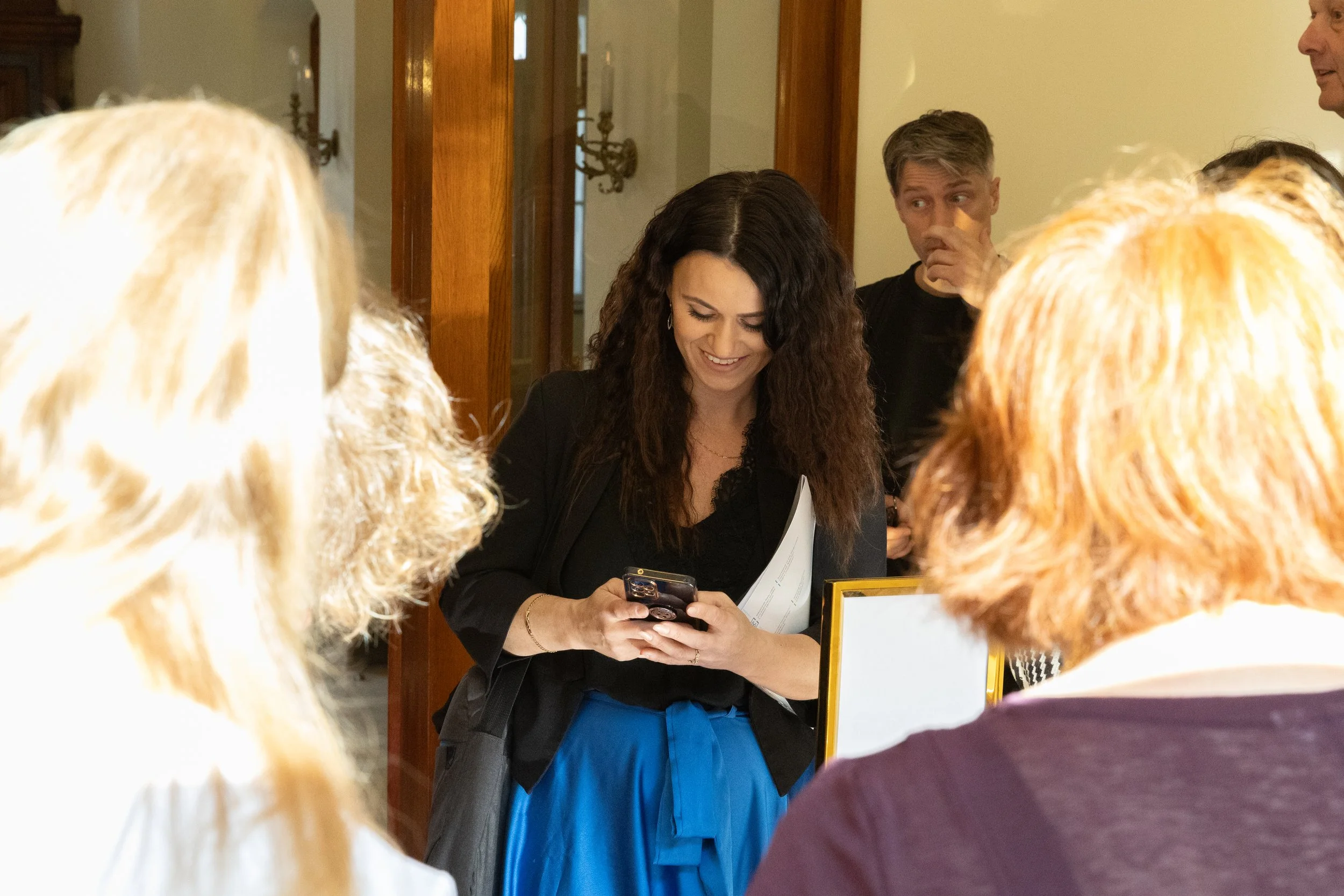 A group of people gathered around a woman with dark, curly hair, who is smiling and looking at her phone. Other individuals near her are partially visible, with some showing their hair and shoulders. The setting appears to be indoors, possibly at a s