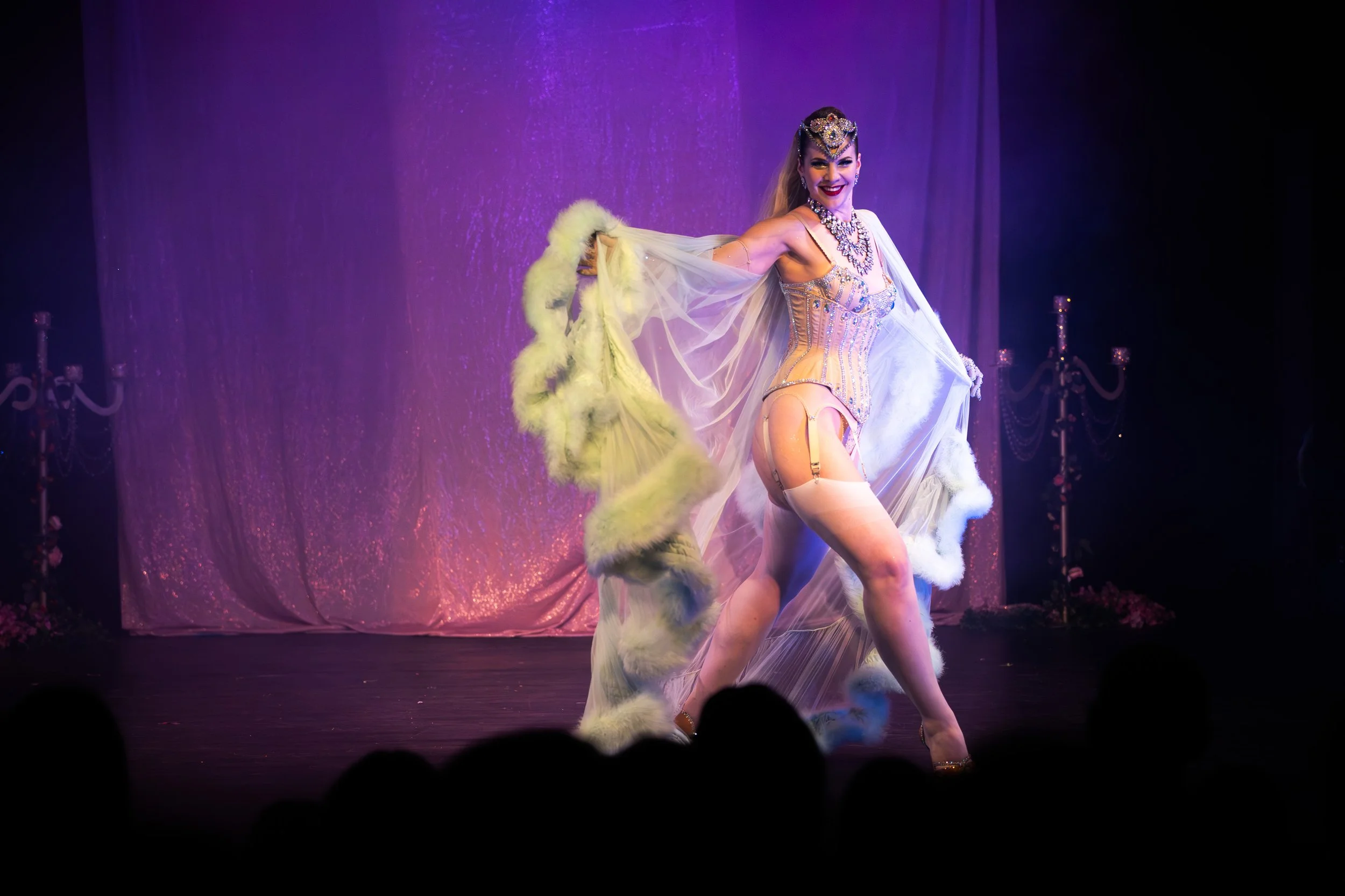A performer on stage in an elaborate costume, posing confidently with a purple-lit background and audience silhouettes in the foreground.