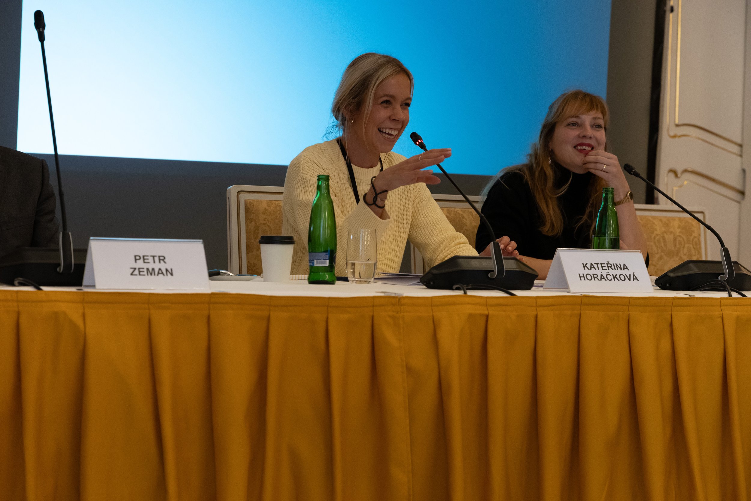 Two women sitting at a conference table with microphones, smiling and engaging in a discussion, with nameplates reading PETR ZEMAN and KATERINA HORACKOVA.