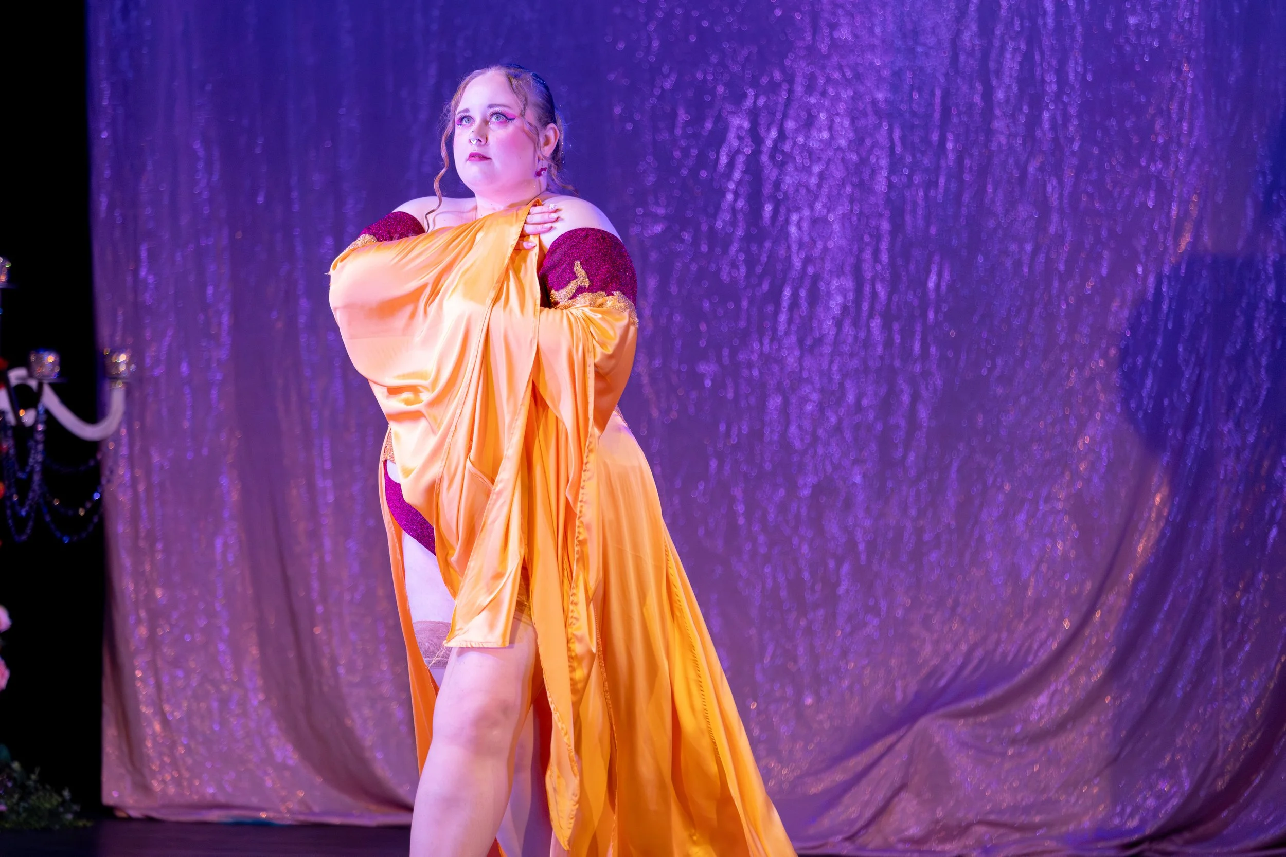 Person performing on stage with purple glittery curtain background, dressed in an elaborate costume with yellow fabric and maroon accents.