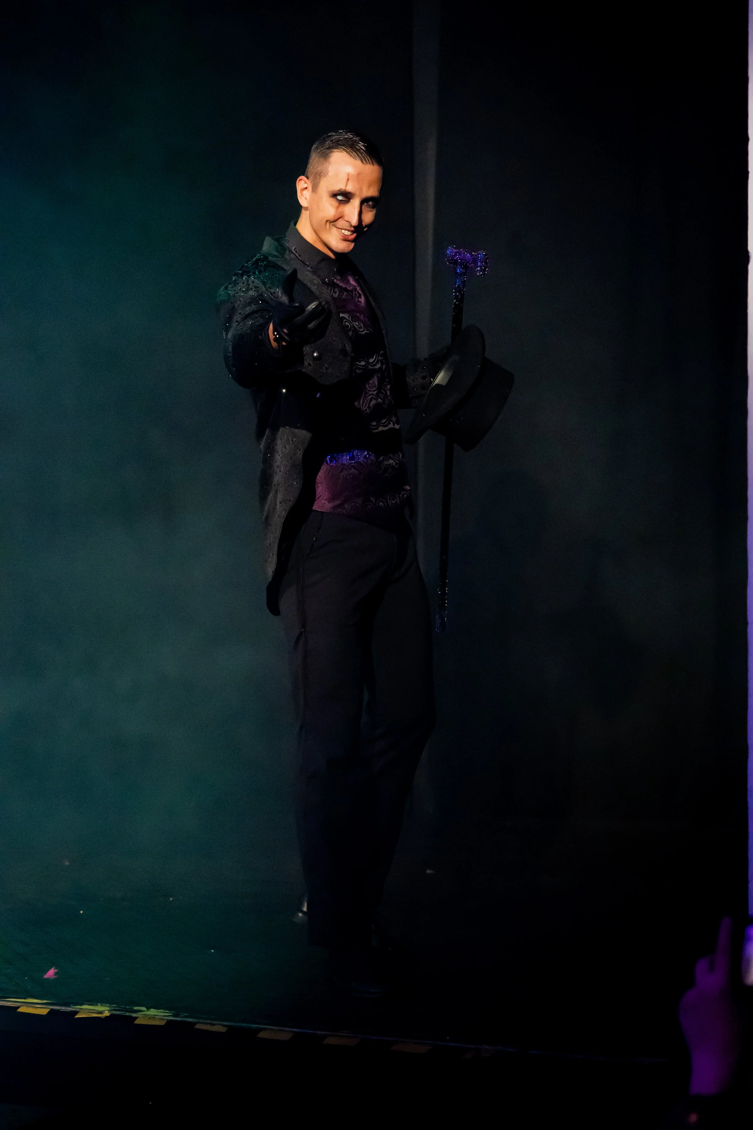 Person dressed as a magician, holding a hat with a magic wand on top, smiling and pointing towards the camera in a dark setting