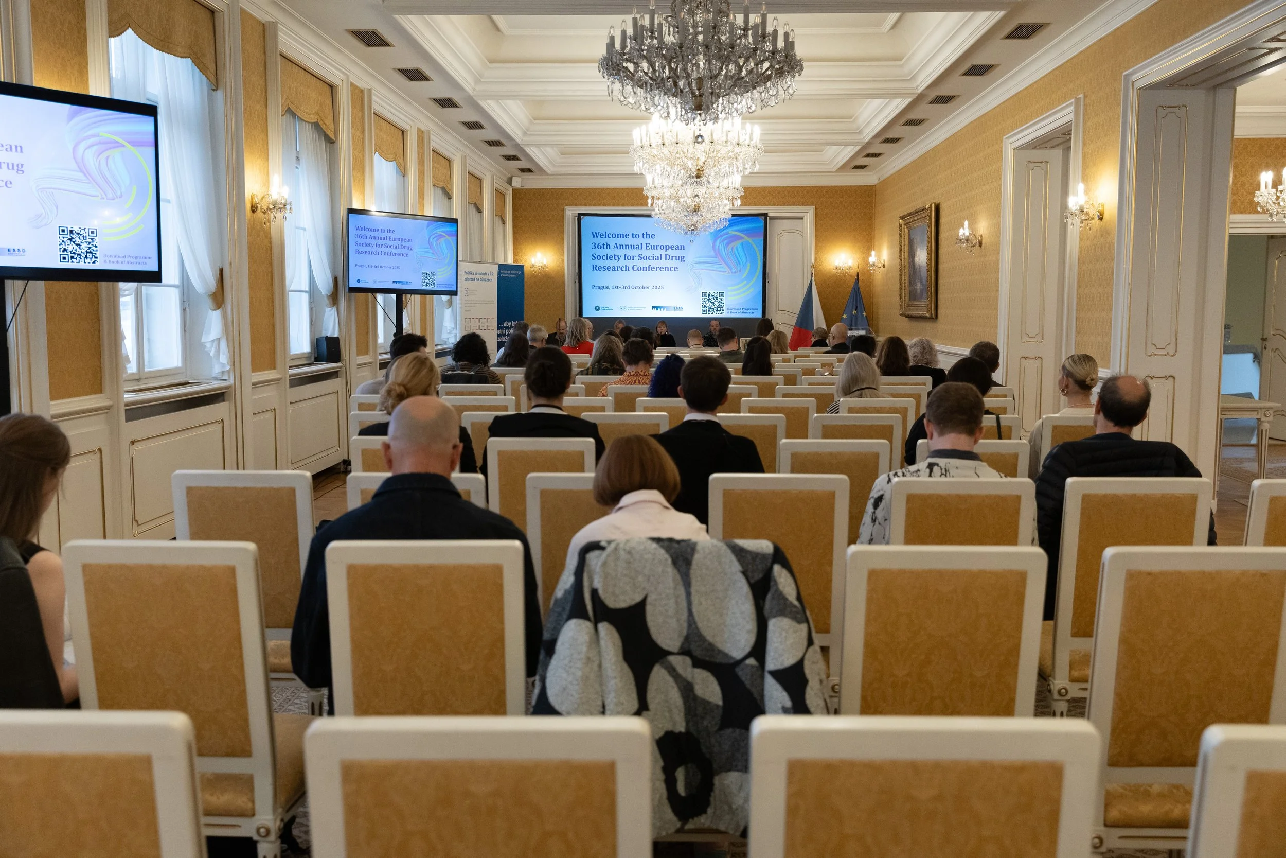 Conference room with attendees seated in rows facing a large screen. The room has ornate decor, chandeliers, tall windows with draped curtains, and flags at the front.