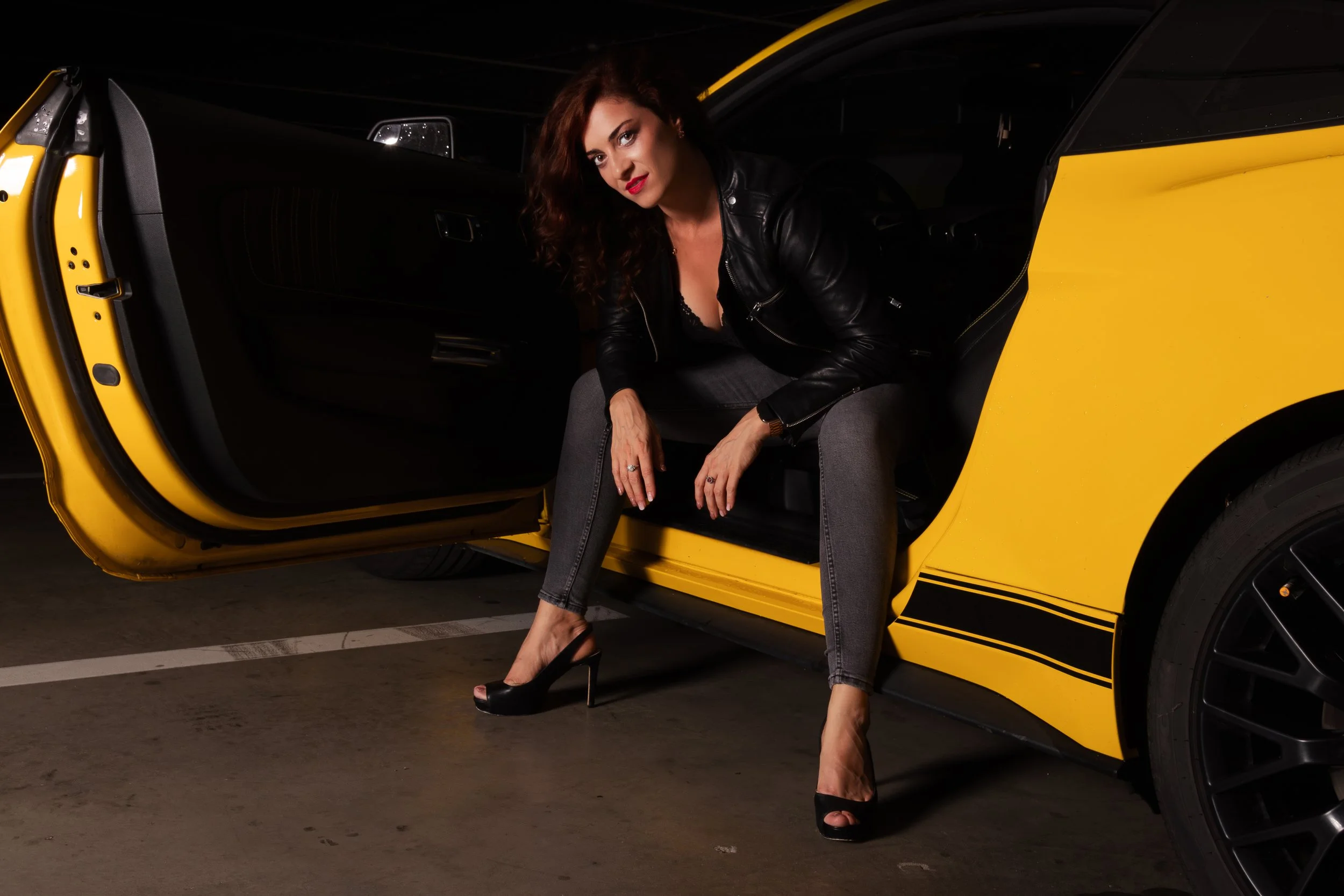 A woman sitting in a yellow sports car with the door open, wearing a black leather jacket, gray jeans, and black high heels.