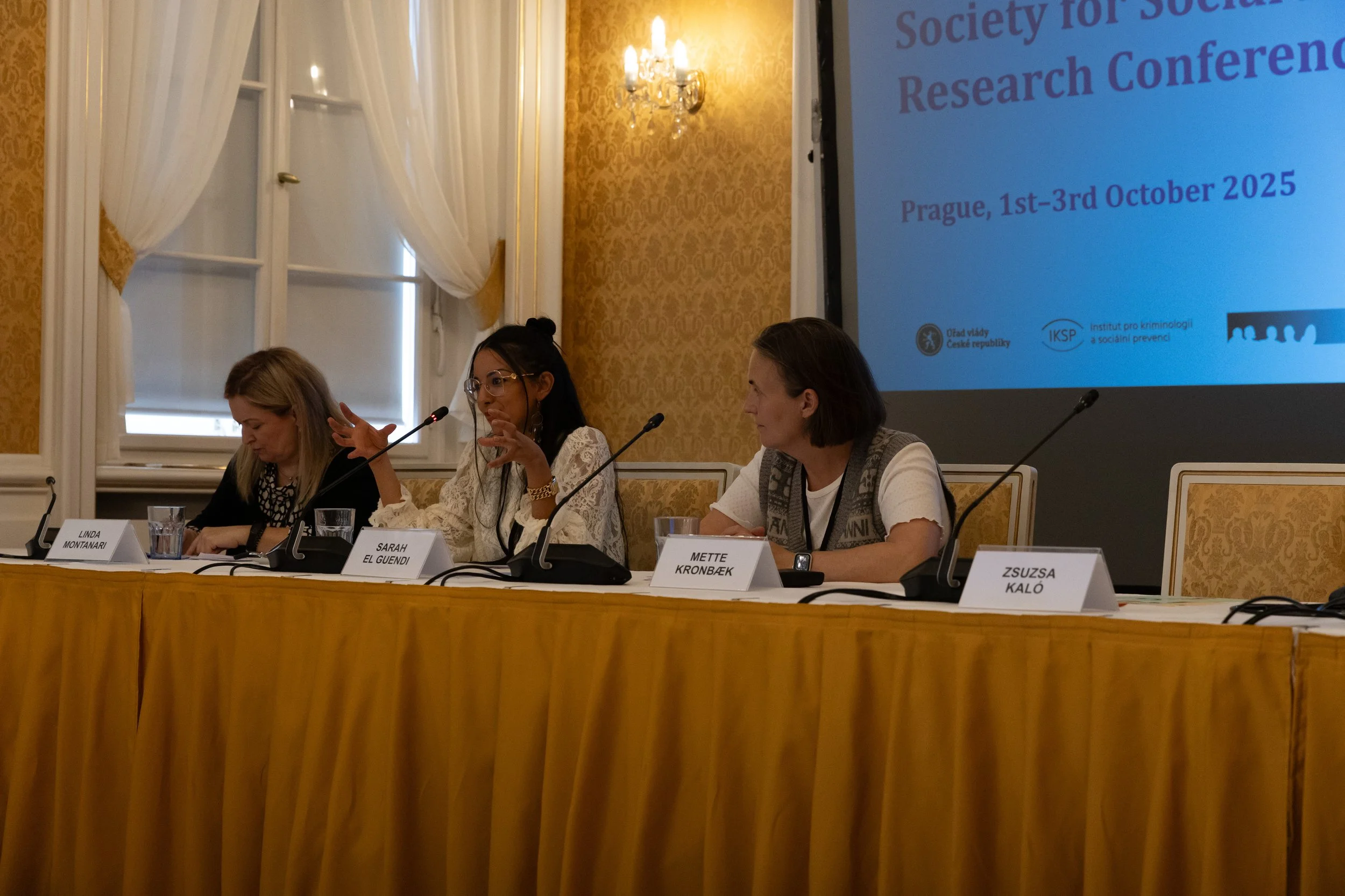 Four women sitting at a conference table with microphones, nameplates, and glasses of water, in a room with ornate wallpaper, large windows with curtains, and a large presentation screen displaying information about a social research conference in Pr