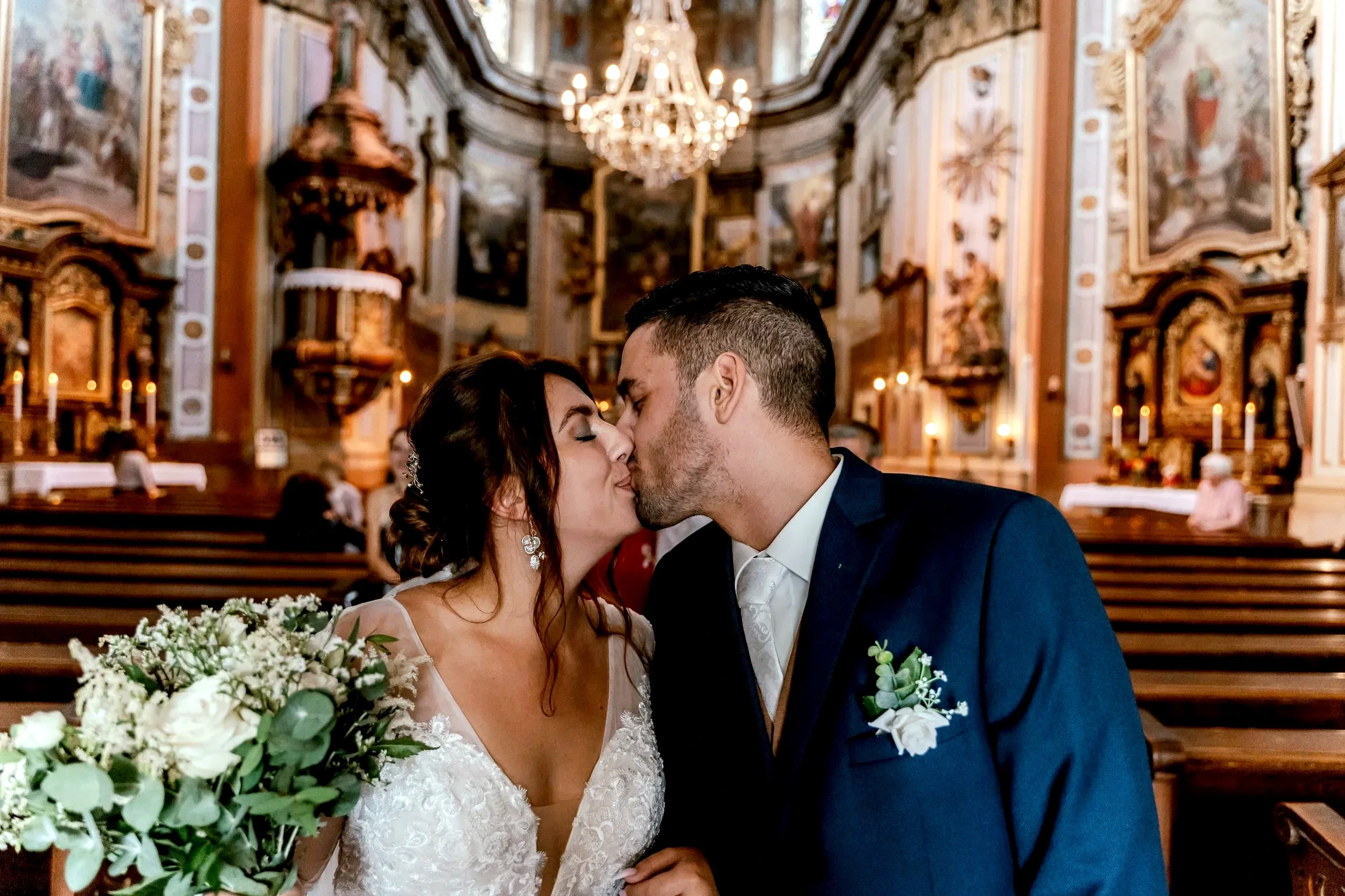 A bride and groom sharing a kiss inside a church with ornate decorations, a chandelier, and religious paintings.