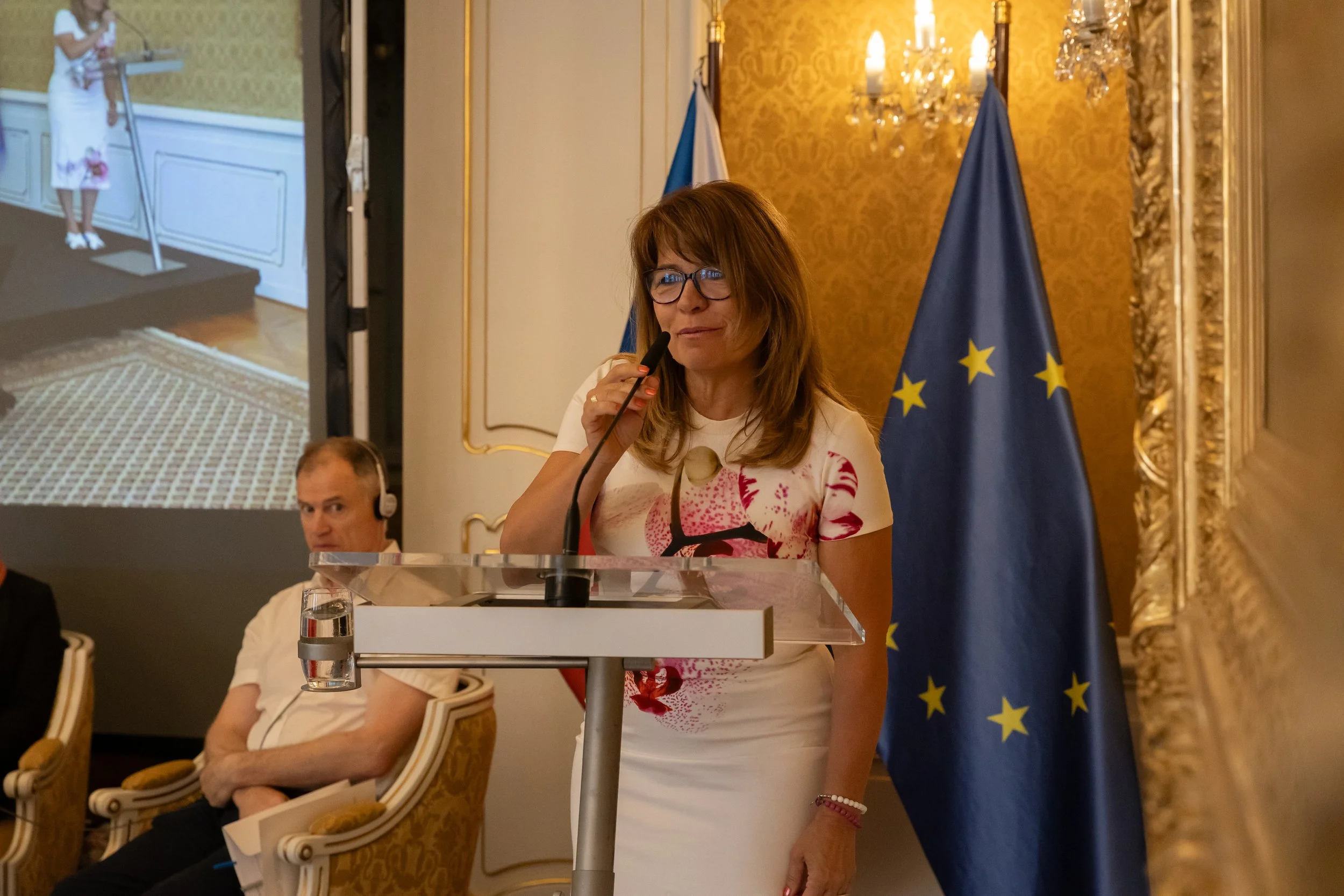 A woman with brown hair and glasses stands at a podium, speaking into a microphone, with European Union flags in the background. Two men are seated behind her in a formal room with ornate gold and white decor.