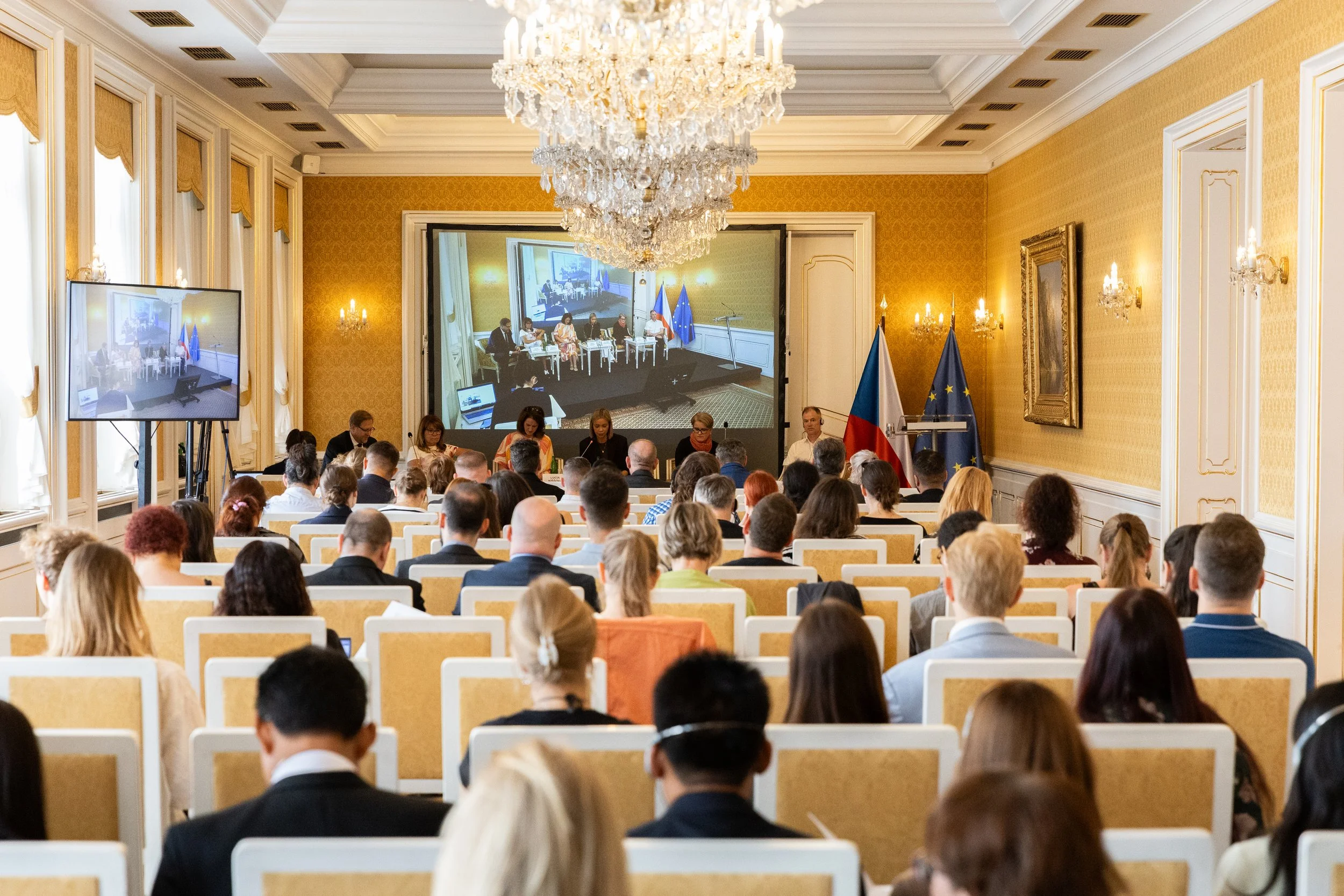 A conference room filled with seated attendees facing a panel of speakers at a long table. The room has a large crystal chandelier, gold-colored walls, and flags of the Czech Republic and the European Union. There are two large screens displaying the