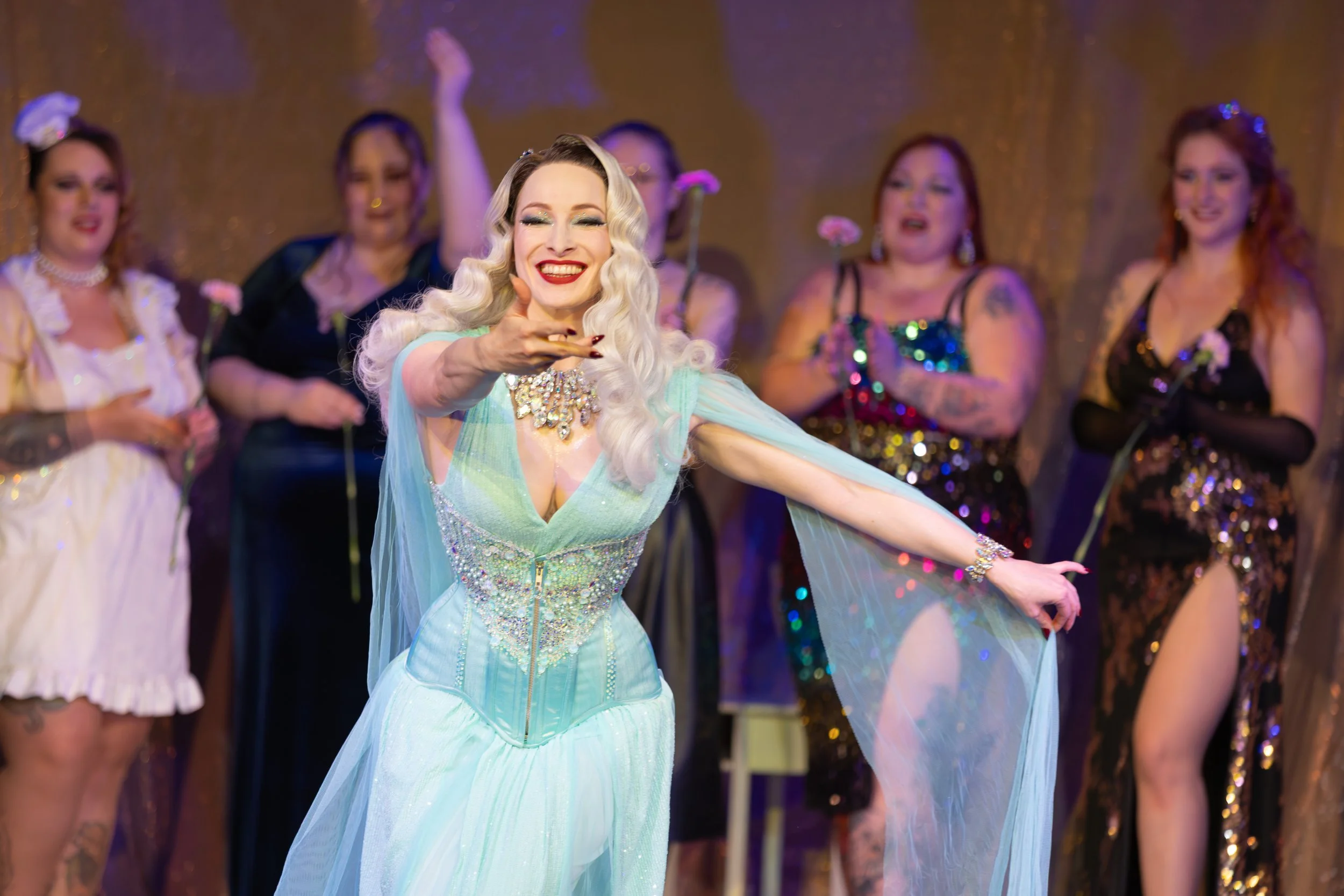 A woman with platinum blonde hair and red lipstick is smiling and blowing a kiss on stage, wearing an elegant light blue gown with rhinestones and sheer cape, surrounded by other women in glamorous dresses.
