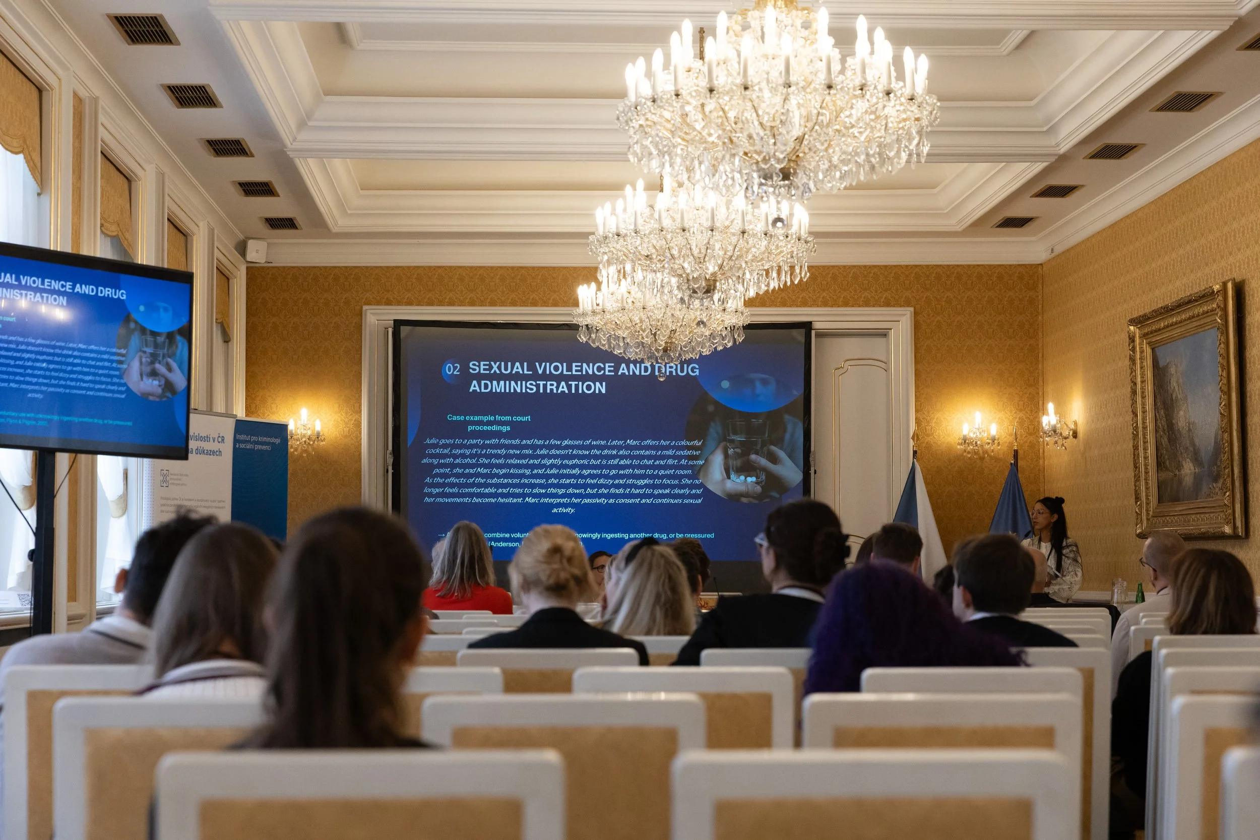 A conference room filled with attendees watching a presentation on sexual violence and drug administration, with a speaker at a podium, large screens displaying the slide, ornate golden walls, chandeliers, and flags.
