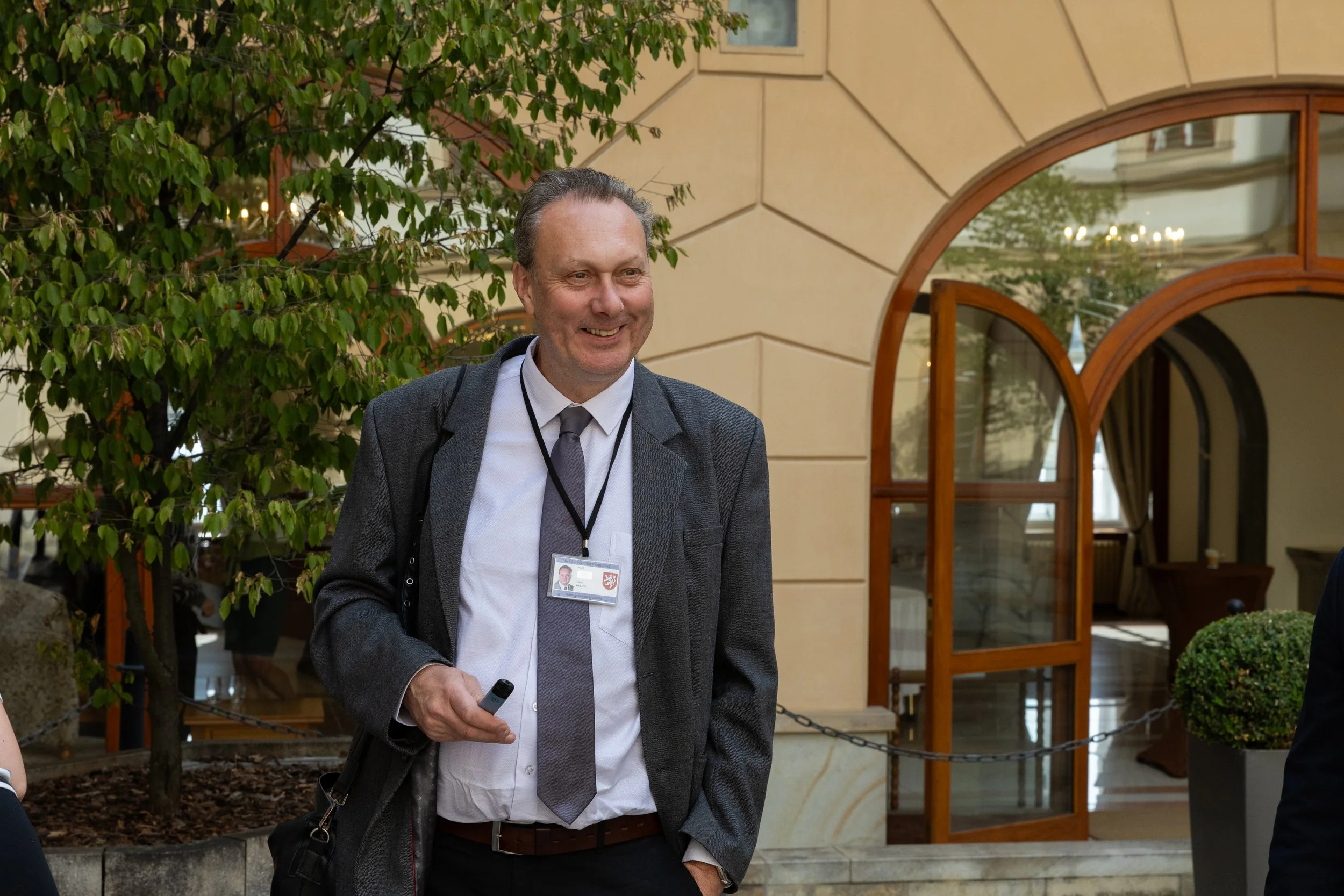 A smiling man in a gray suit and white shirt, holding a device, standing outdoors near a building with arched windows and greenery.
