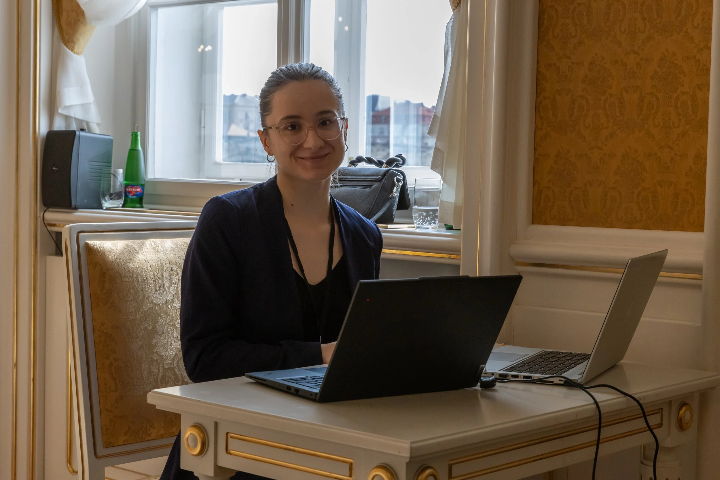 A young woman with glasses sitting at a desk in a well-decorated room, smiling at the camera with two laptops in front of her, a window with city view behind her.