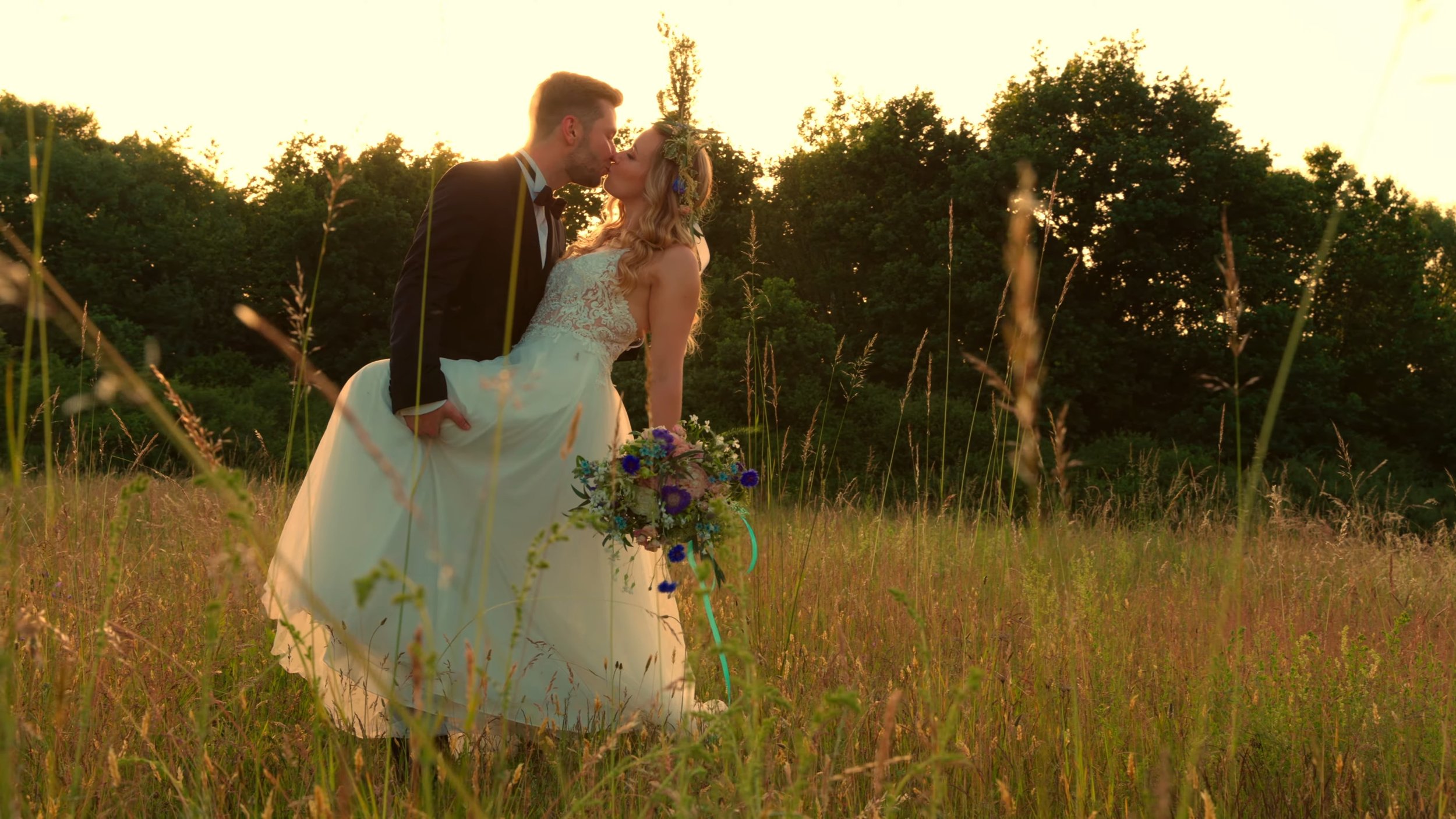 A newlywed couple kissing in a grassy field during sunset, with the groom wearing a tuxedo and the bride in a white wedding dress holding a bouquet of purple and white flowers.