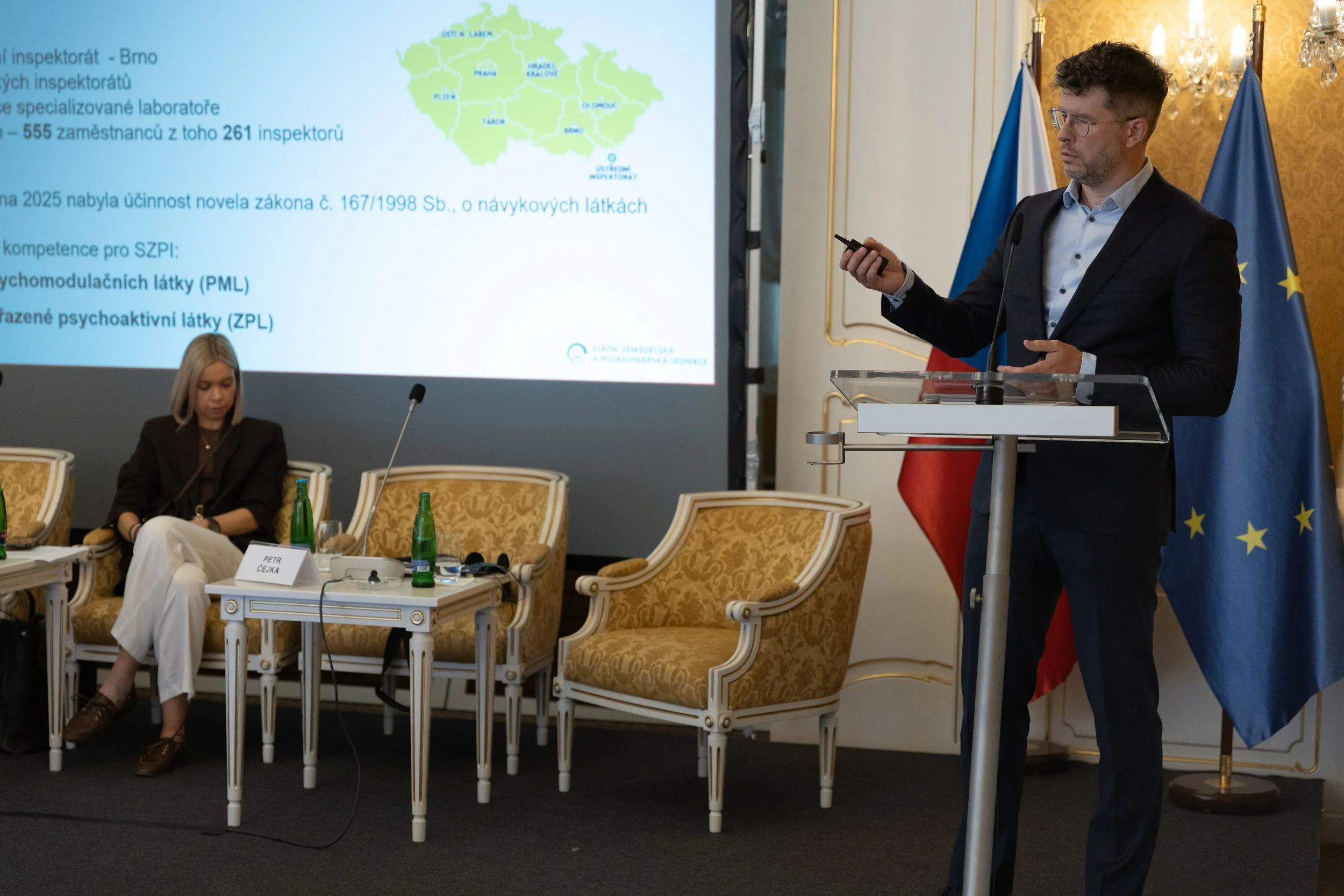 A man in a dark suit and glasses standing at a podium delivering a presentation, with a woman seated to his left and a large screen displaying a map and text in the background. The setting is a formal conference room with gold-patterned wallpaper and