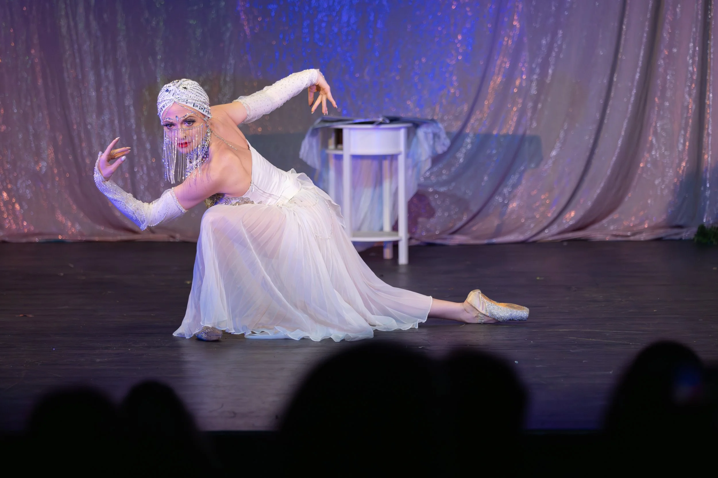 A woman dressed in a white gown with glittery accessories performs a dance on stage. She wears a head wrap and a face veil with beads, and sparkly shoes. The stage has a purple glittery curtain backdrop with a small table covered with fabric in the b
