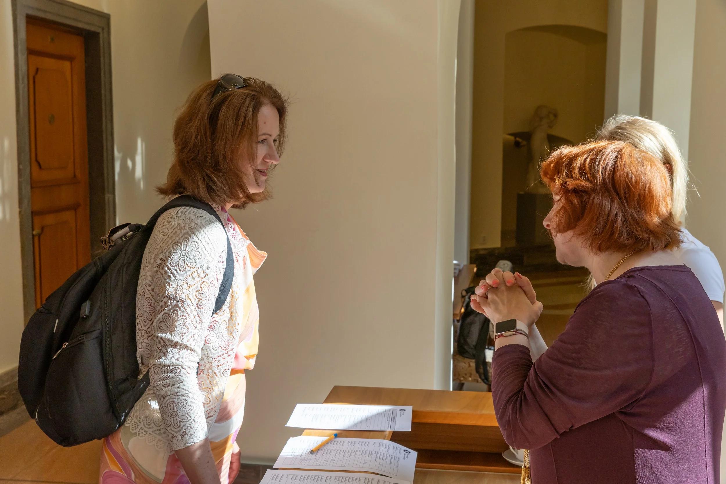 Two women conversing at a reception desk, one with a backpack and the other wearing a smartwatch, with papers and pens on the counter.