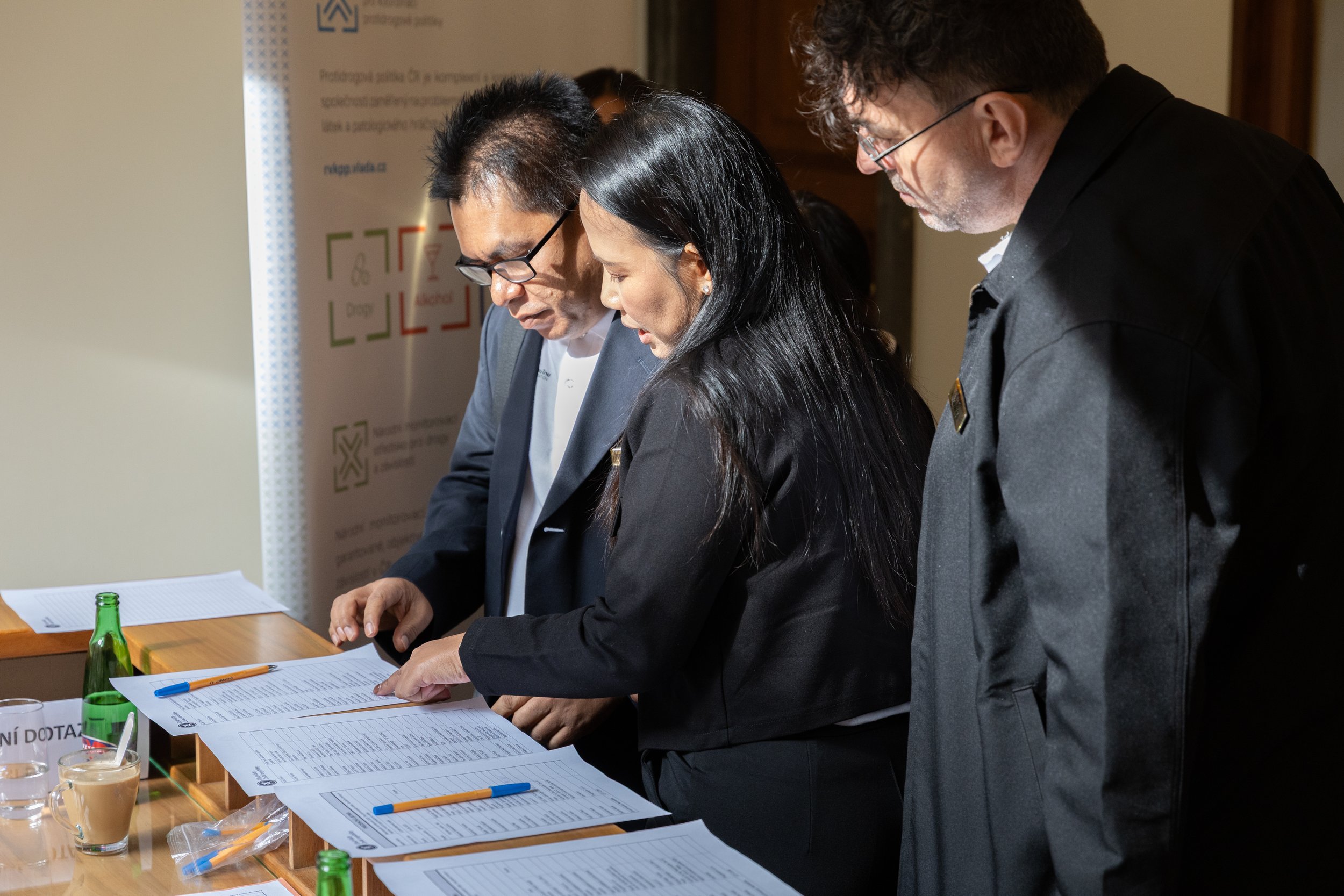 Three people in formal attire are leaning over a table, examining documents with pens on top, and drinks placed nearby.