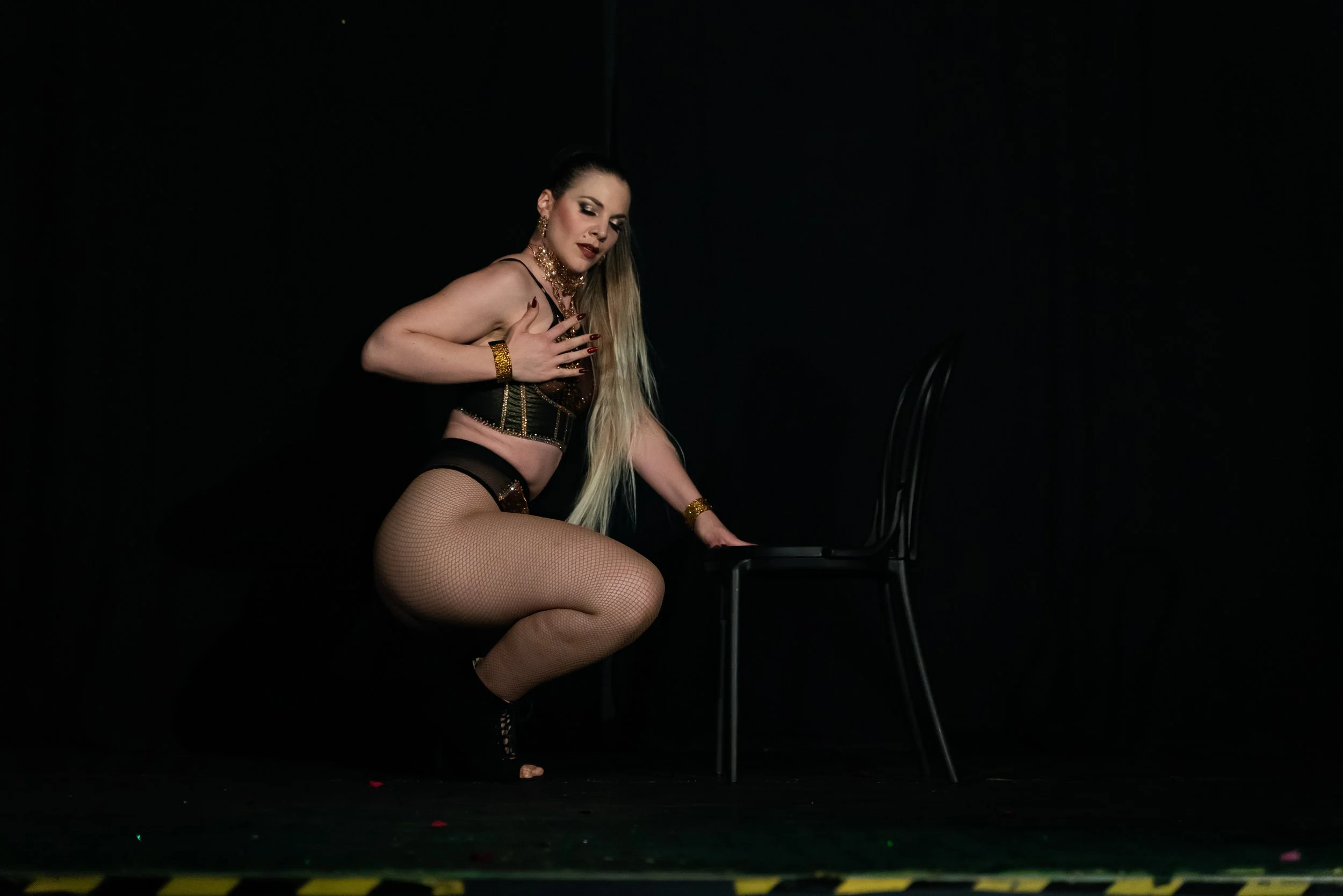 A woman with long blonde hair, wearing fishnet stockings, black heels, and black lingerie, is kneeling on a dark stage next to a black chair, with one hand on the chair and the other touching her chest.