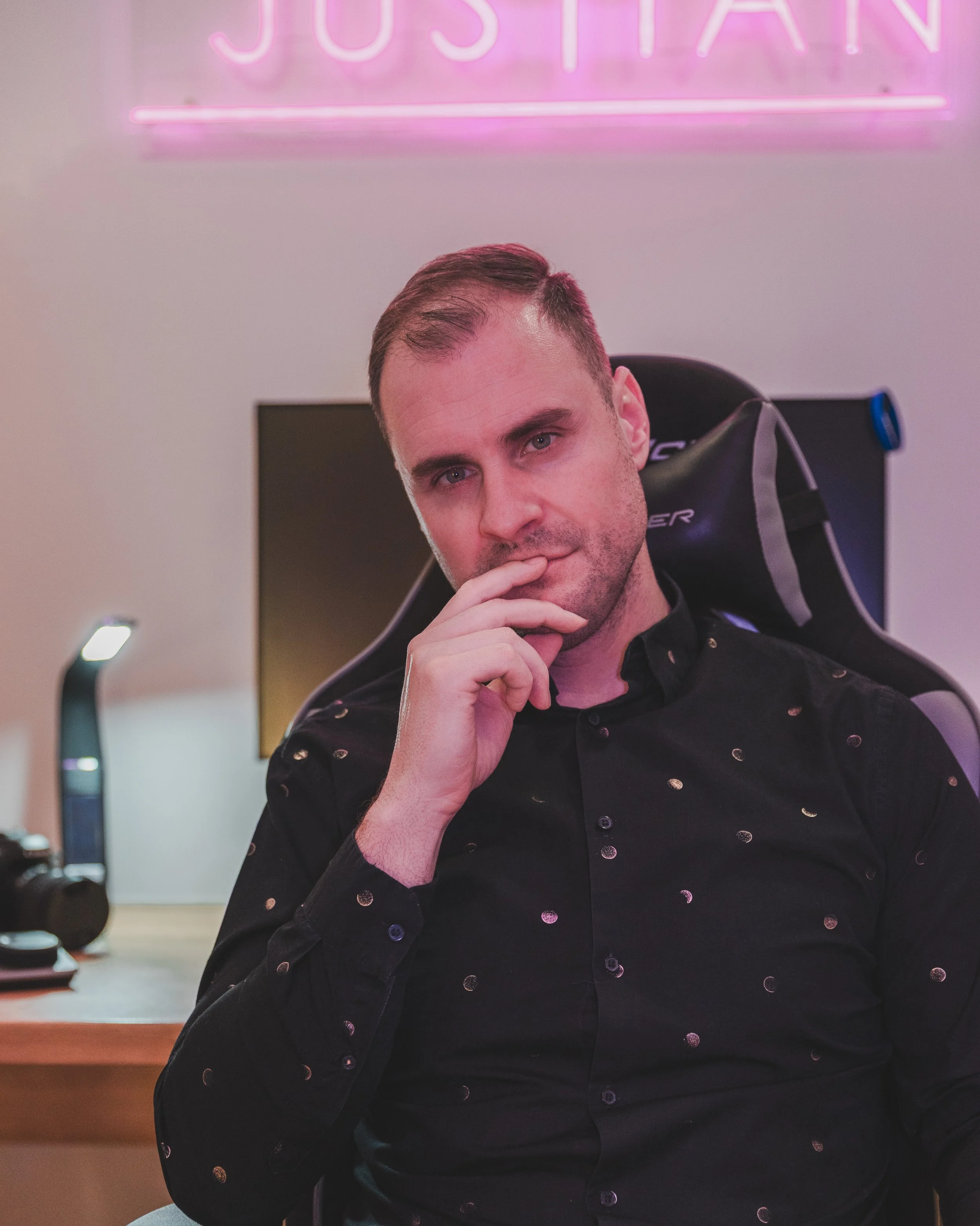 A man sitting in a gaming chair with a hand on his chin, wearing a black shirt with small metallic dots, in a room with a pink neon sign that says 'JOSTIAN' and various tech items on a wooden desk.
