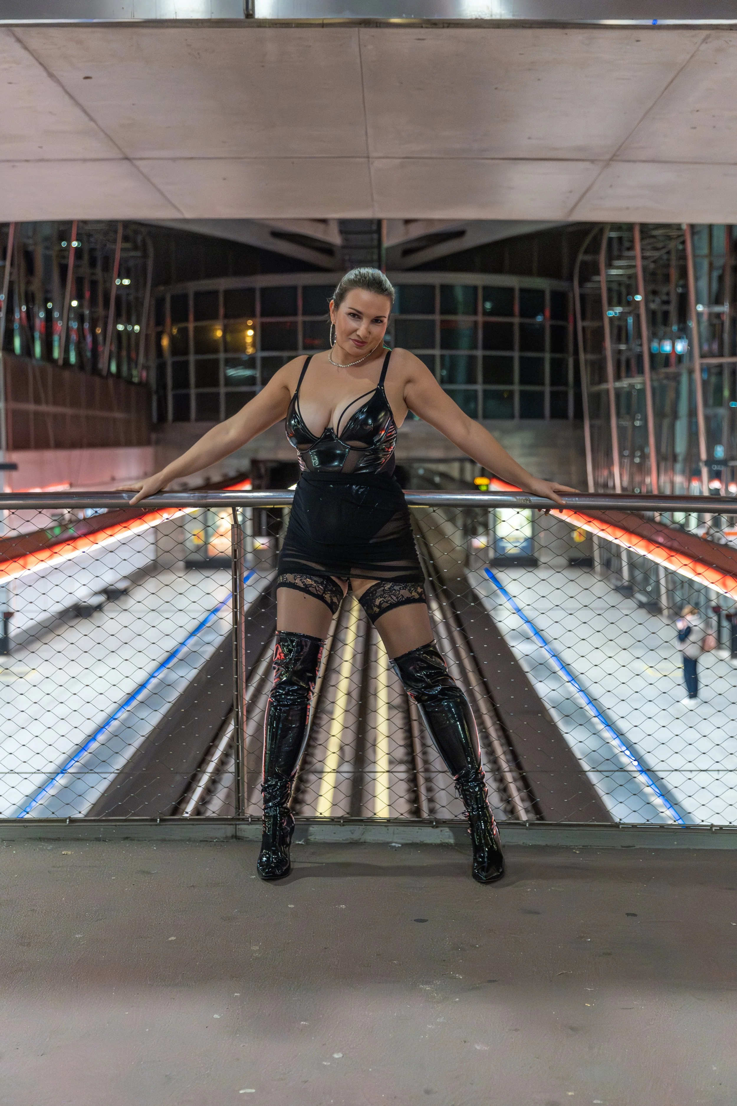 A woman in black latex clothing and thigh-high boots standing on an elevated platform in a modern, brightly lit indoor space with a curved glass roof and illuminated surroundings.