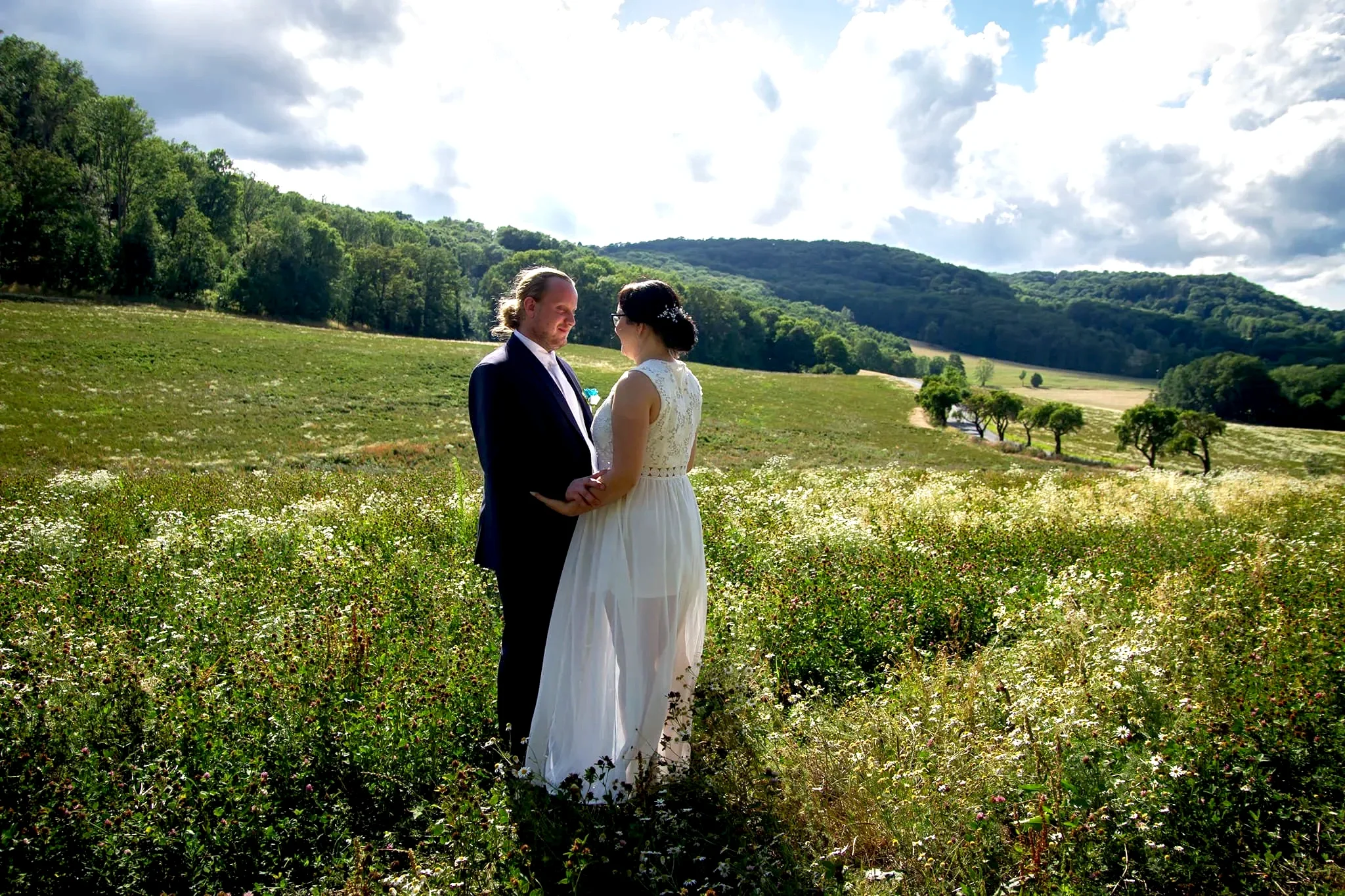 A couple in wedding attire holding hands and facing each other in a lush green field with wildflowers, surrounded by rolling hills and trees under a partly cloudy sky.