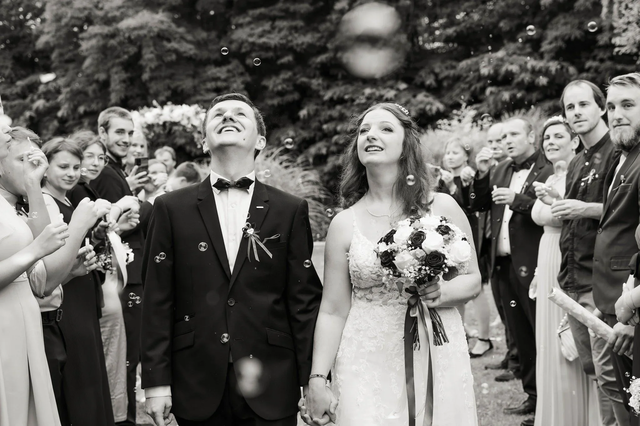 A black and white photo of a newlywed couple holding hands, surrounded by friends and family at their wedding celebration, with bubbles floating in the air.
