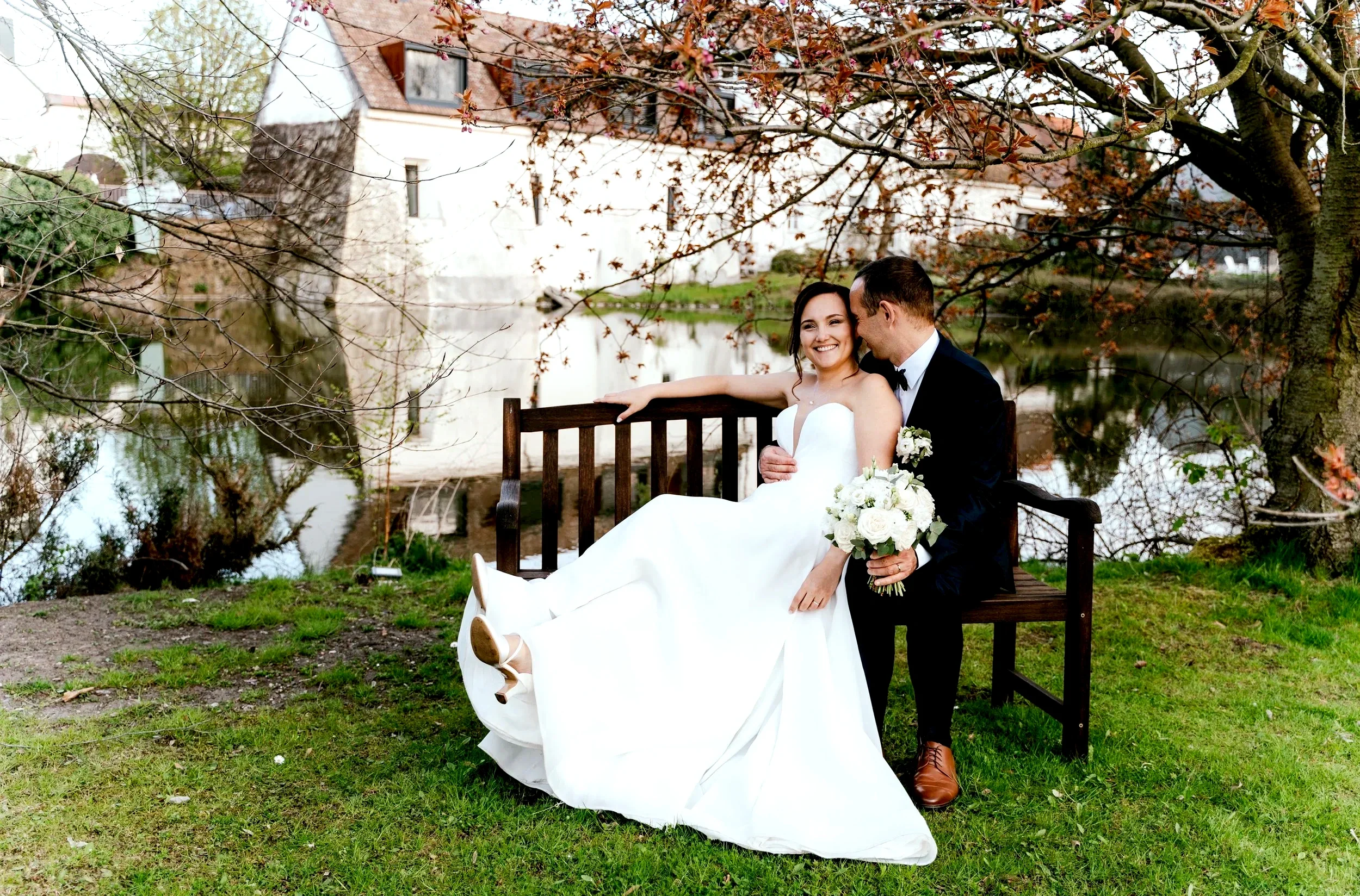 A bride and groom sitting on a wooden bench next to a lake under a tree with pink blossoms. The bride is wearing a white wedding dress and holding a bouquet of white roses, while the groom is in a black tuxedo with a bow tie, leaning close and kissing her.