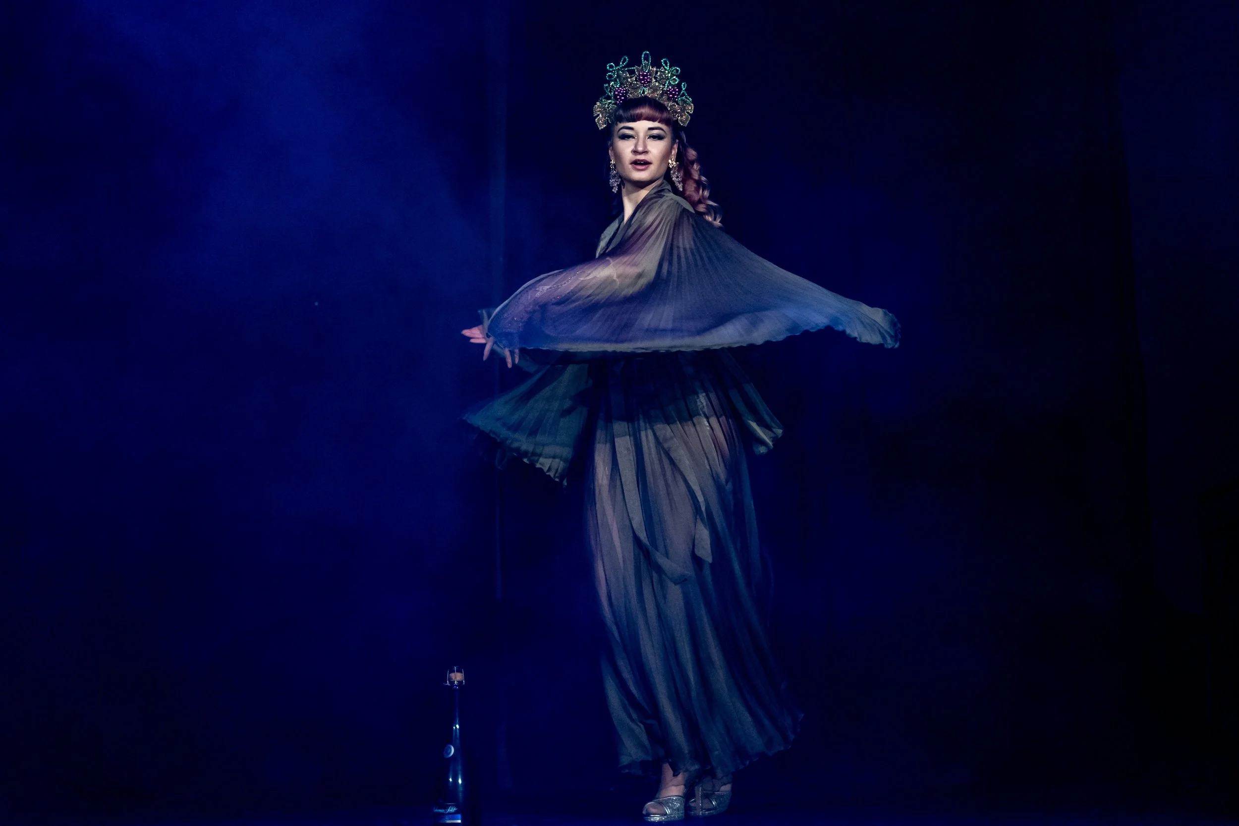 A woman performing a dance, wearing a flowing, layered black dress and a decorative crown, on a dark stage with blue lighting.