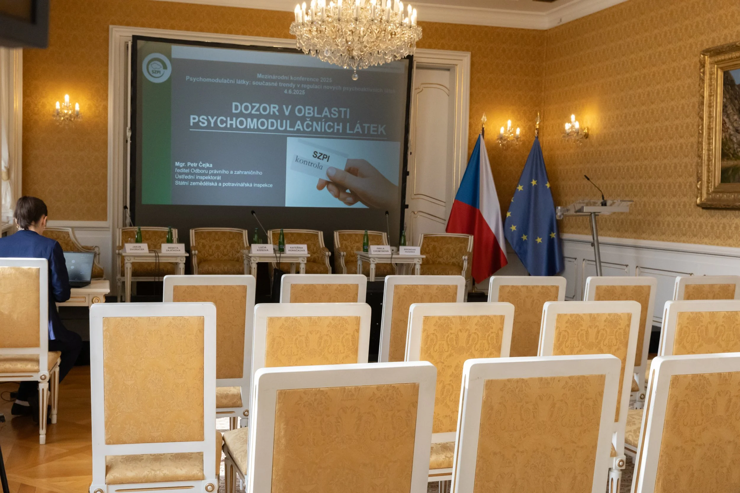 Empty conference room with gold patterned walls, chandelier, and flags of Czech Republic and European Union, set up for a presentation with a large screen displaying a title slide and a woman working on a laptop.
