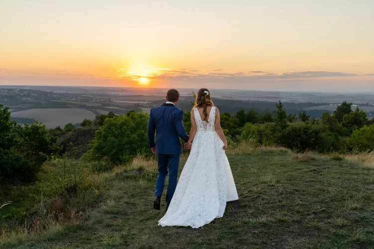 A bride and groom holding hands and walking on a grassy hilltop during sunset, with a scenic landscape of fields and trees in the background.