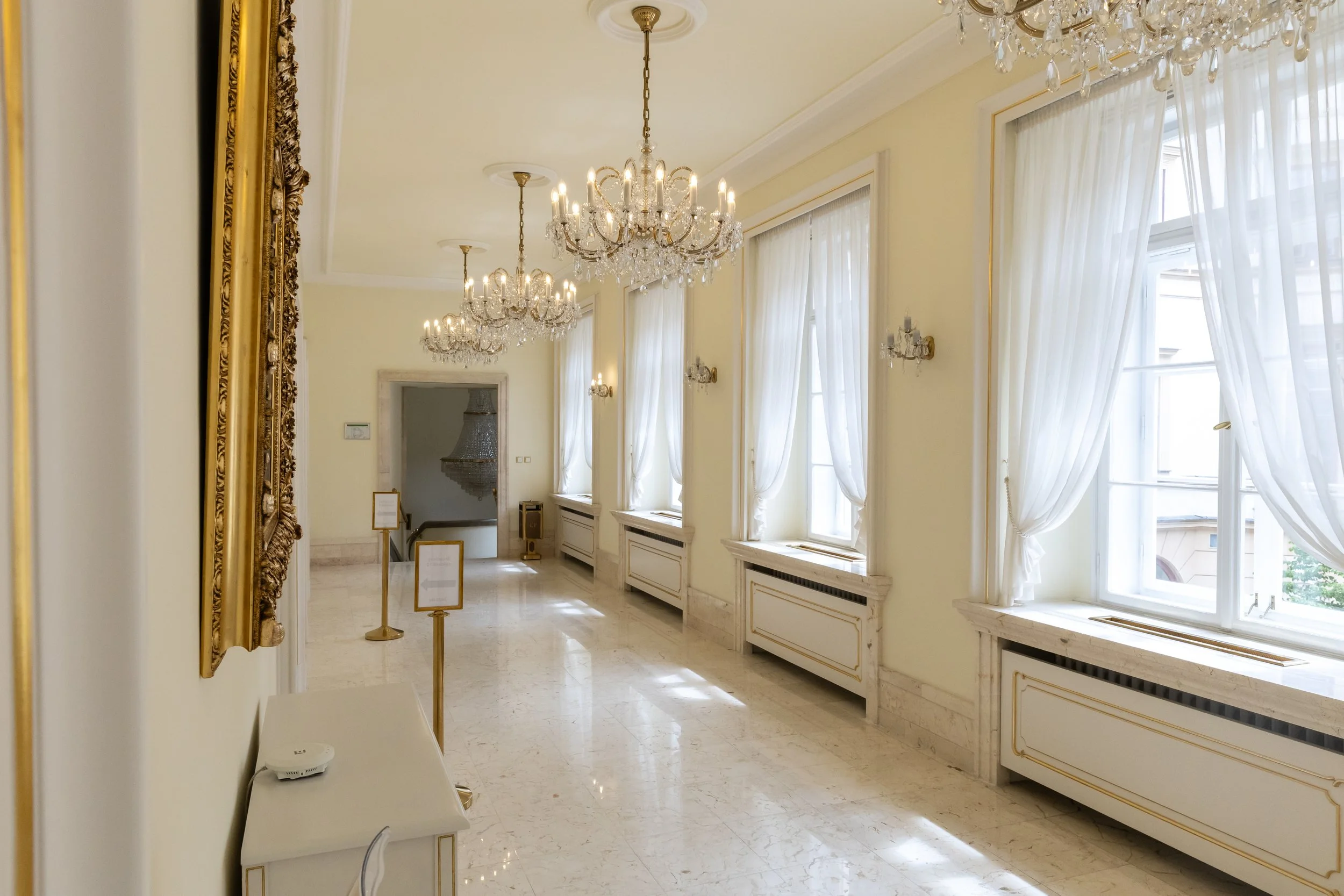 Elegant hallway with large windows, white curtains, marble floors, and ornate chandeliers.