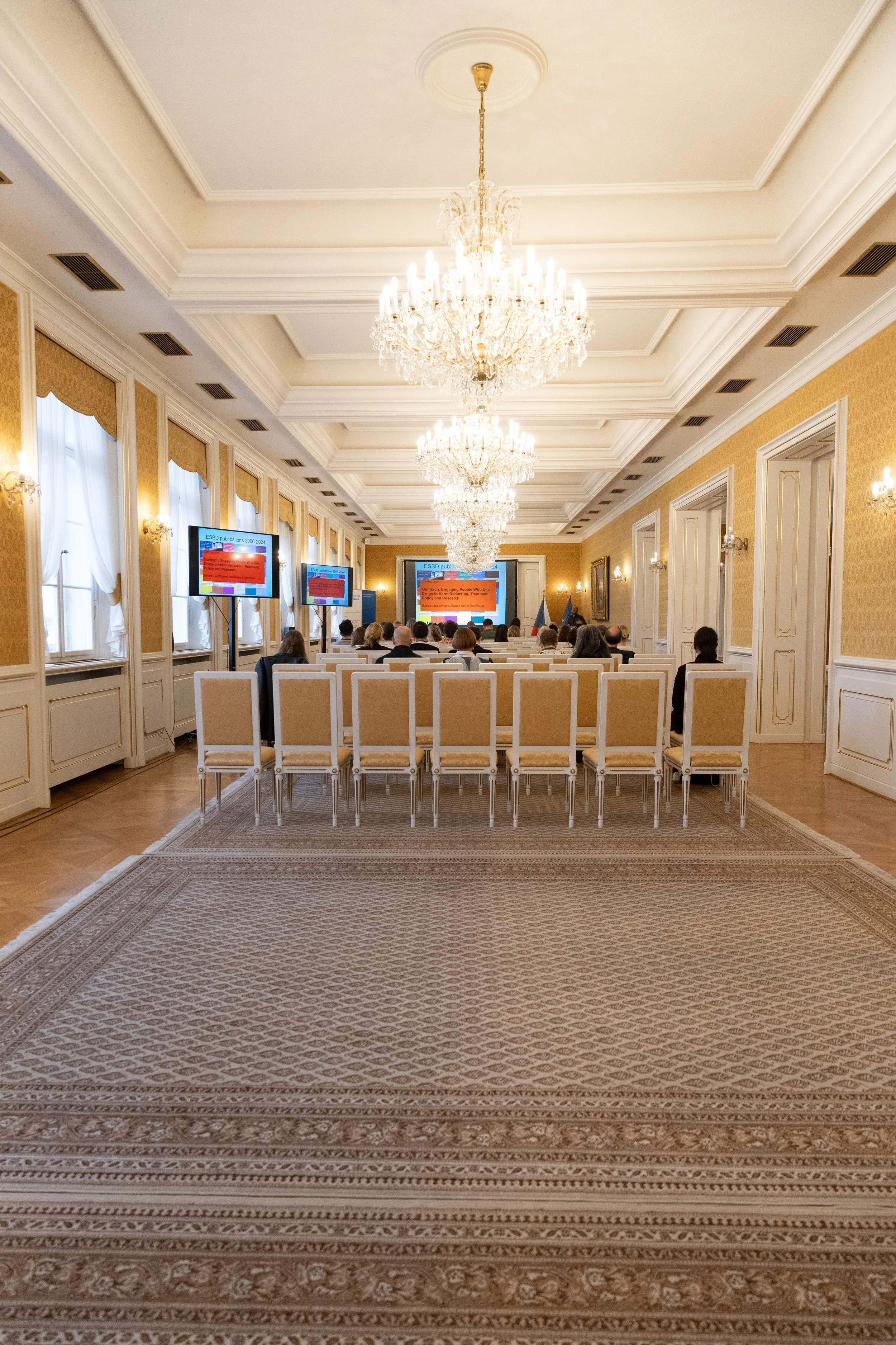 A formal conference room with yellow wallpaper, white trim, chandeliers, large windows with curtains, and a carpeted floor. Attendees sit in rows of chairs facing presentation screens at the front.