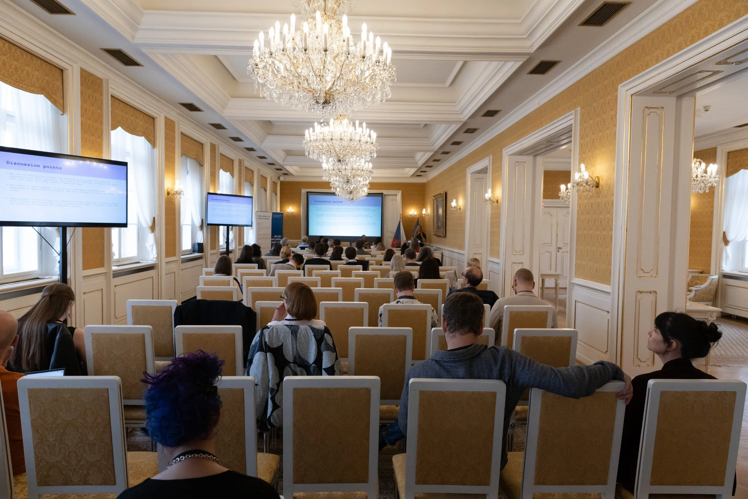 Conference room with chandeliers, attendees seated in rows, and presentation screens at the front.