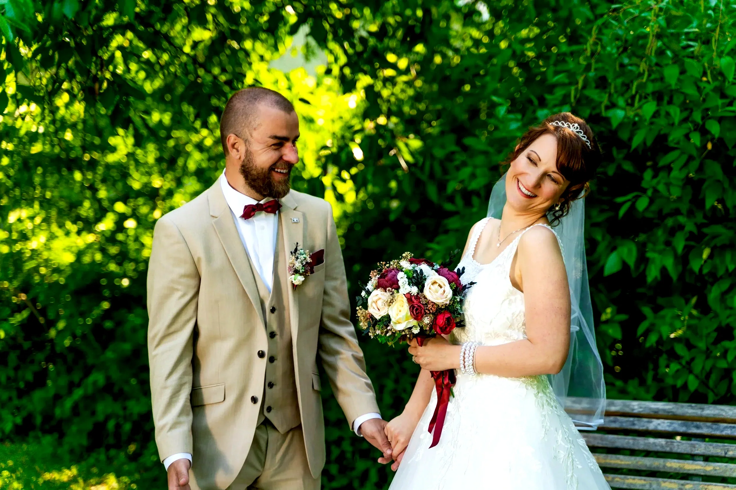 A newlywed couple holding hands in a park with lush green trees in the background. The groom is wearing a beige suit with a red bow tie, and the bride is in a white wedding dress with a veil and holding a bouquet of flowers.