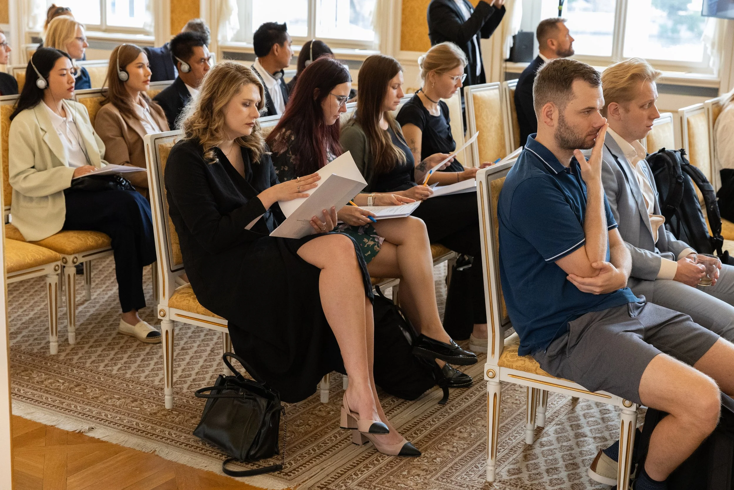 Attendees sitting in a conference room, listening to a speaker at the front, some taking notes and others reading documents.