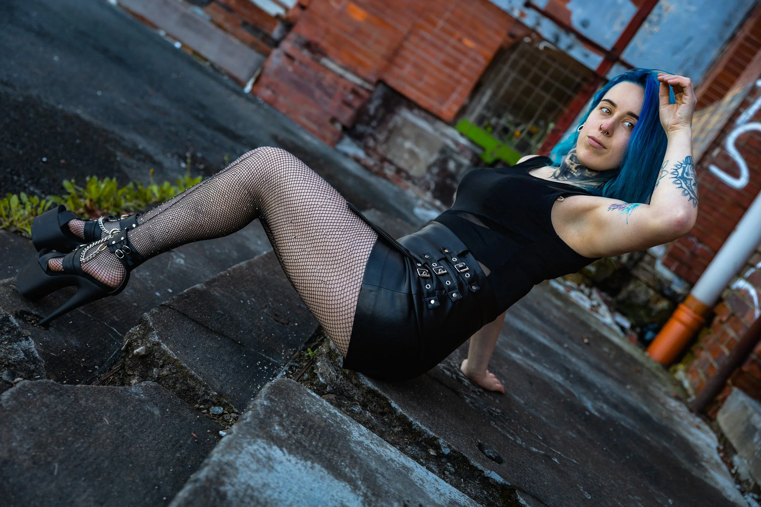 A woman with blue hair, tattoos, wearing fishnet stockings, a black dress, and high heels is seated on a city sidewalk with her legs extended and one hand resting on the ground, against a background of brick walls and graffiti.
