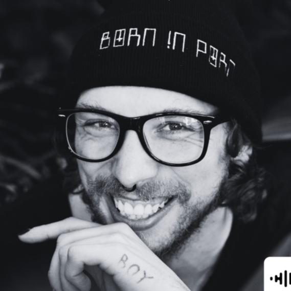 Smiling young man with glasses, wearing a beanie that says 'Born in Portugal', and a tattoo on his hand that says 'BOY'
