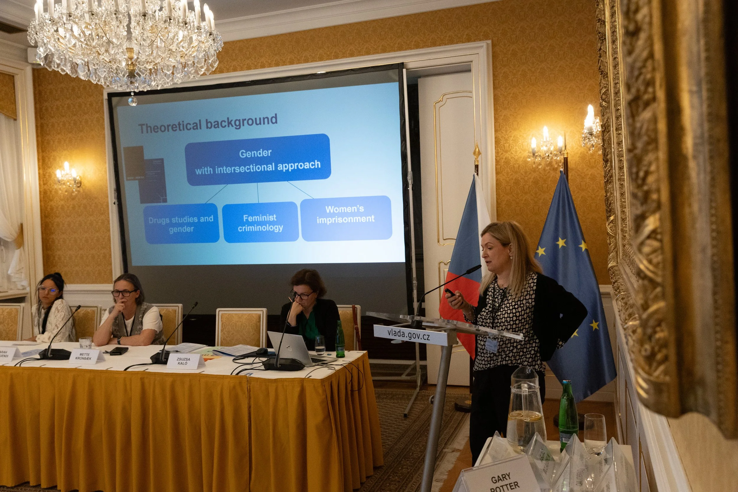 A woman is giving a presentation at a conference with a large screen displaying a slide titled 'Theoretical background' that includes a flowchart with topics such as 'Gender with intersectional approach,' 'Drugs studies and gender,' 'Feminist crimino