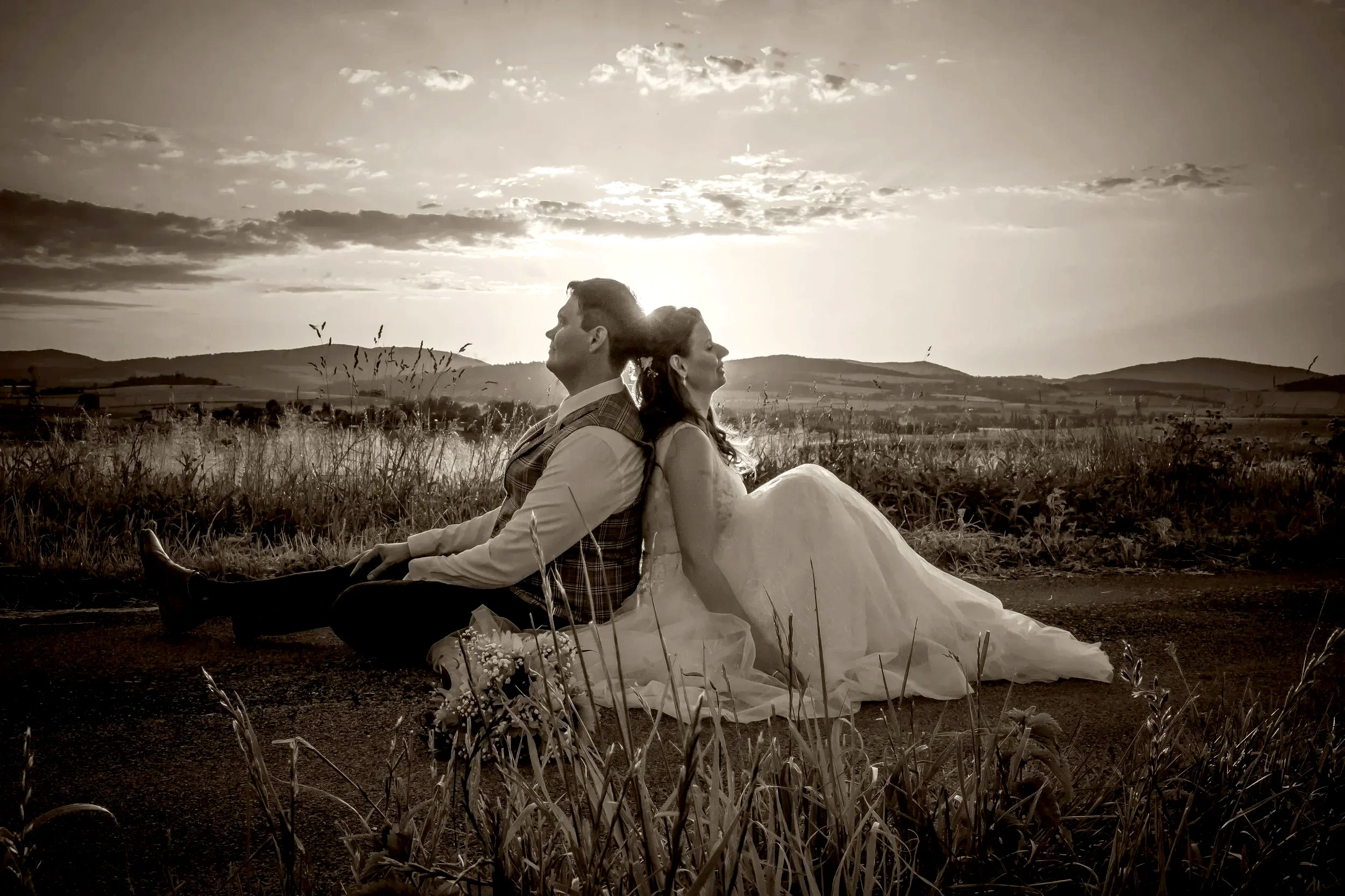 A couple in wedding attire sitting back-to-back on a rural path with tall grass in foreground during sunset or sunrise, with rolling hills in the distance, in a sepia tone.