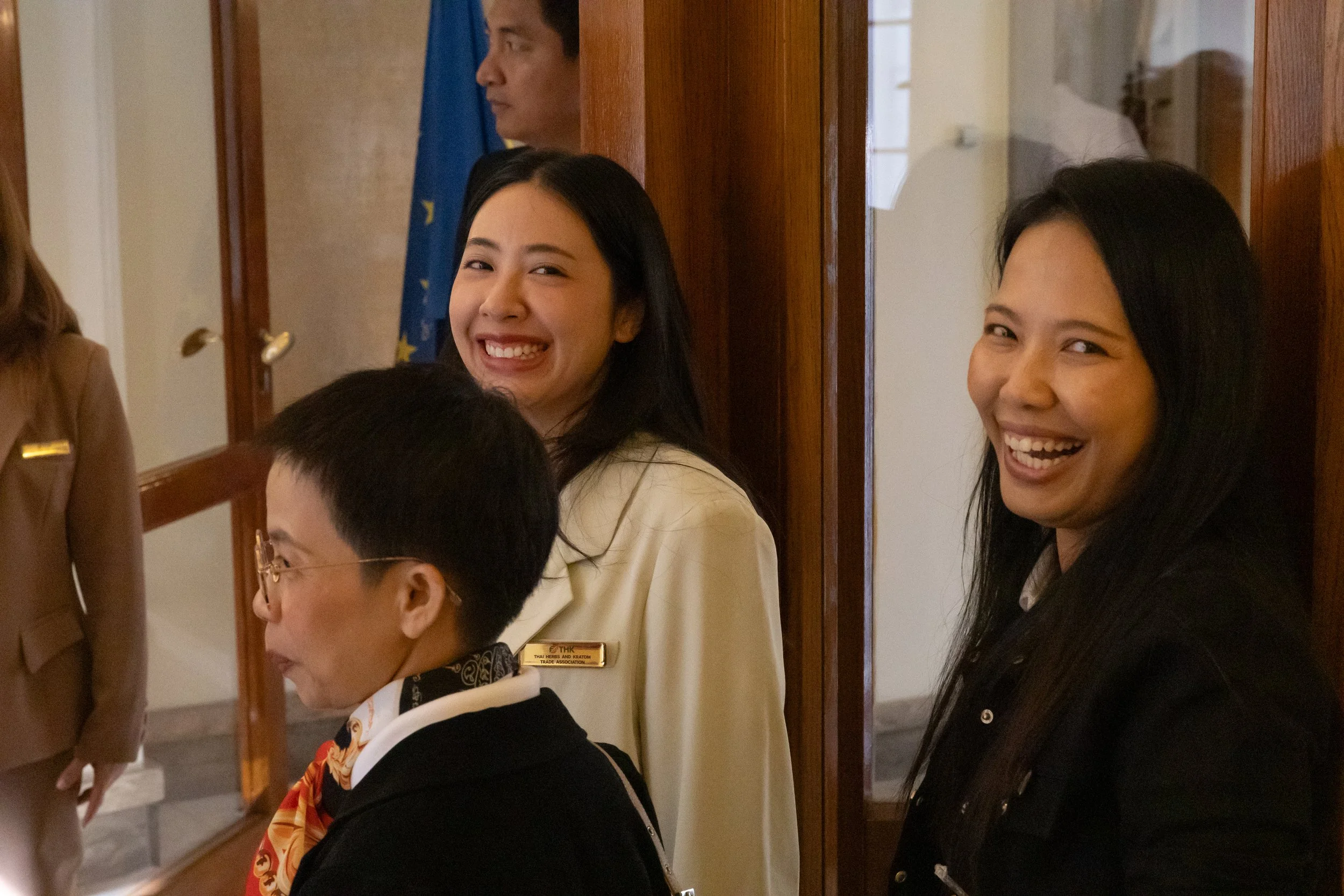 Three women smiling and laughing indoors, two women facing the camera and a woman in glasses looking away, with a woman in uniform nearby and a person in the background.