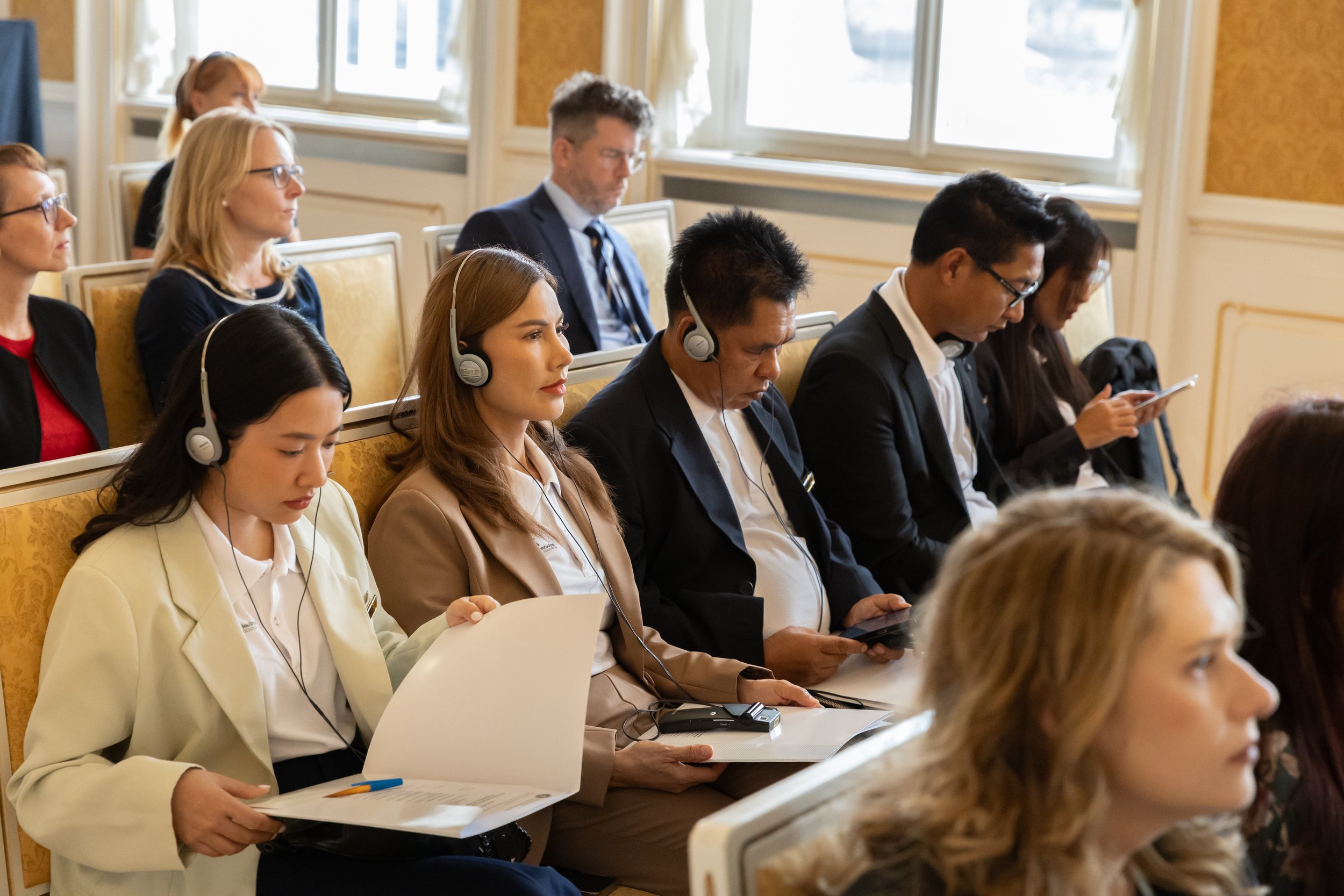 Group of people attending a conference or seminar, sitting in rows, some wearing headsets, some looking at documents or mobile devices, in a well-lit room with large windows.