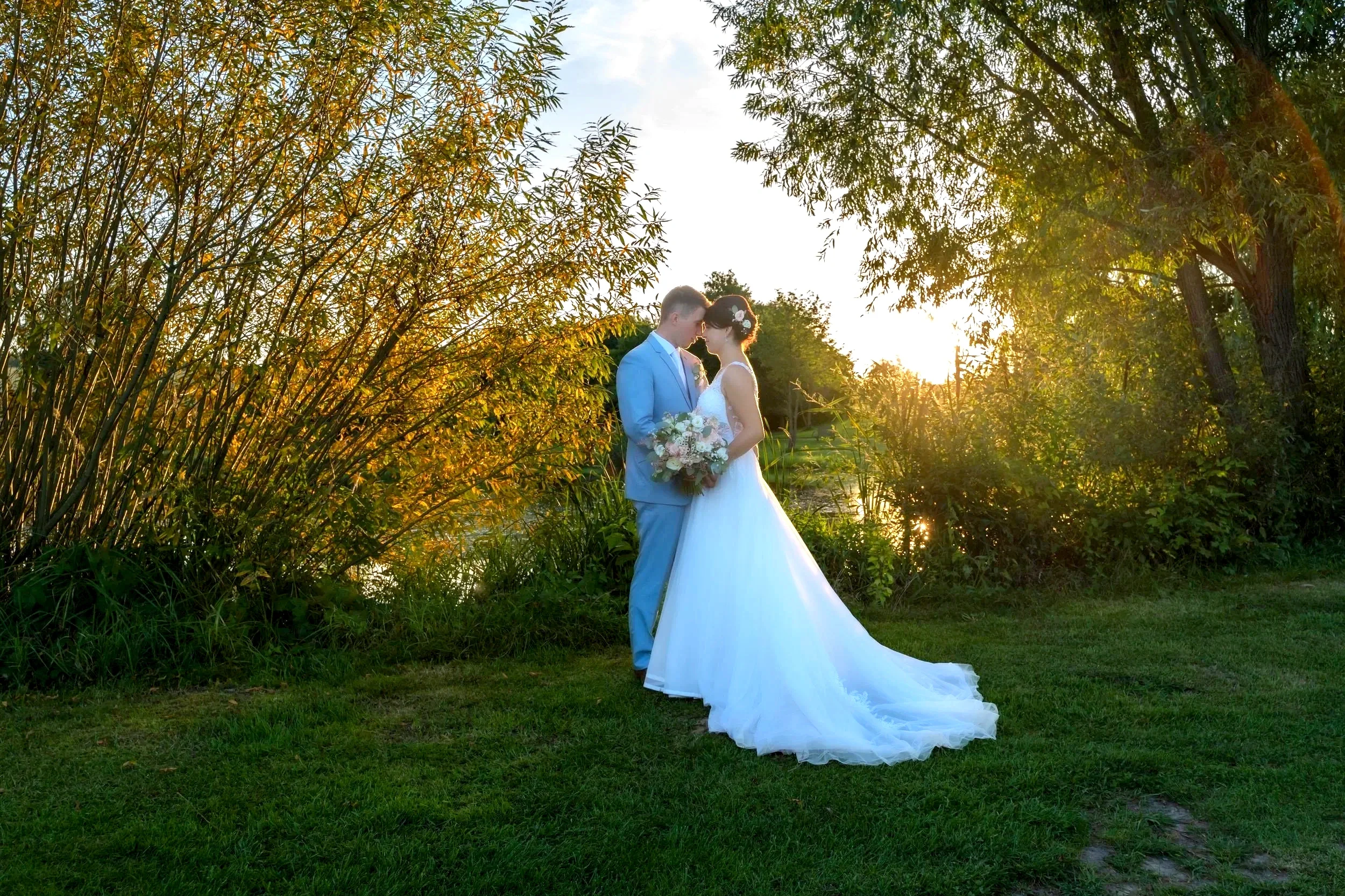 A bride and groom share an intimate moment outdoors during sunset, standing close with foreheads touching, near green bushes and trees, with water in the background.